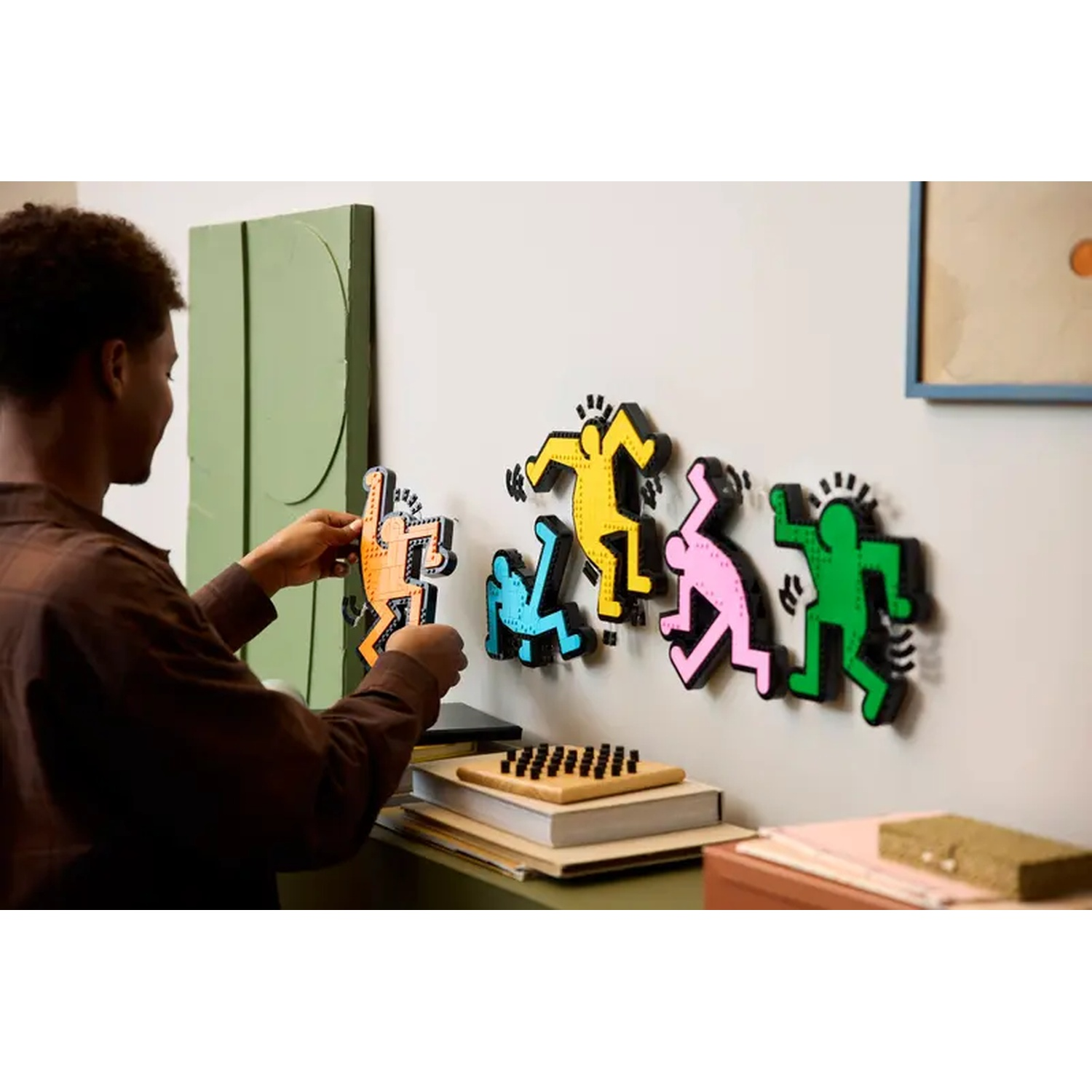 A person hanging art. Colorful stick figures are on the wall. A green abstract art piece and other objects are visible.
