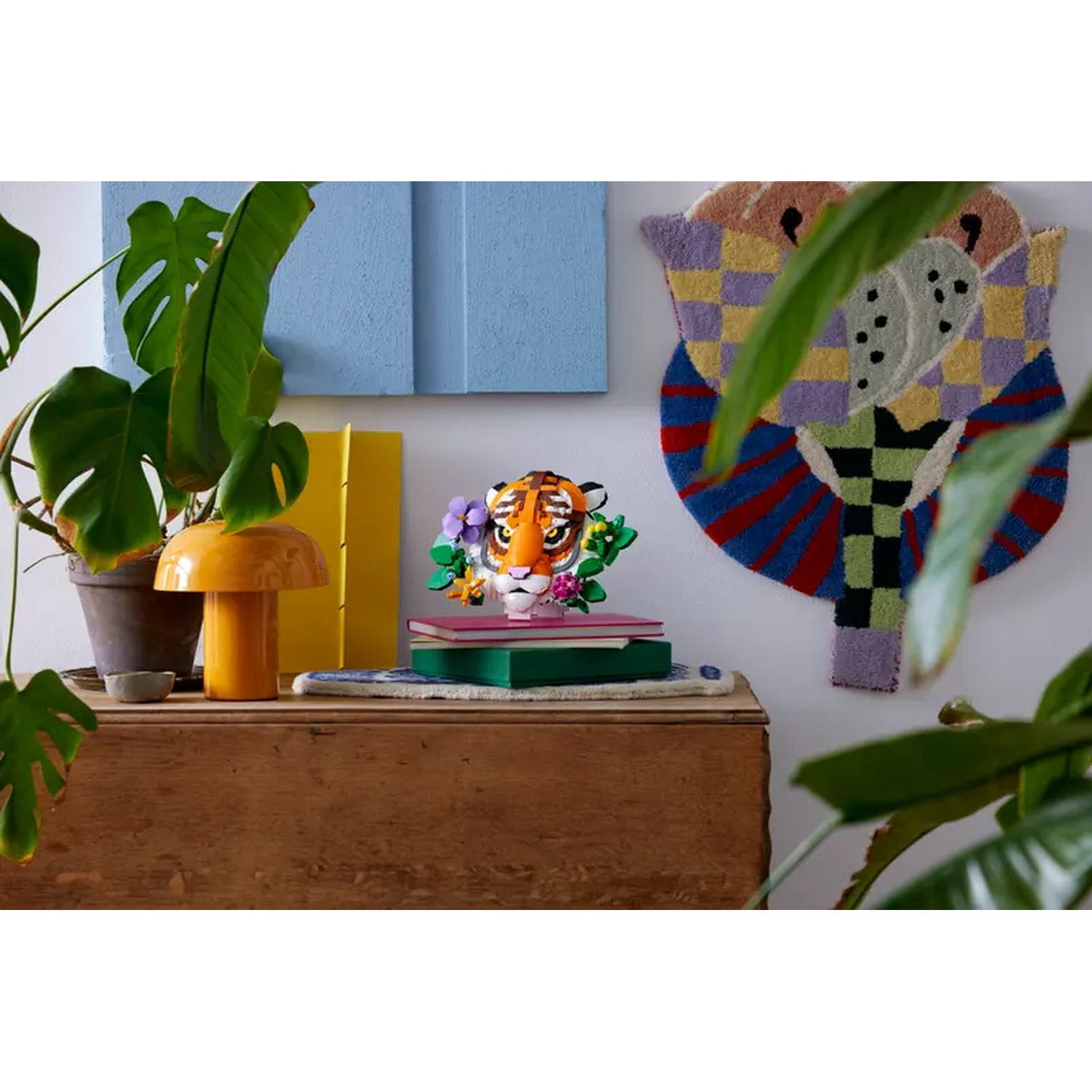 A tiger head sculpture sits on books atop a wooden chest. A yellow lamp and green plants are nearby. A colorful rug hangs on the wall.