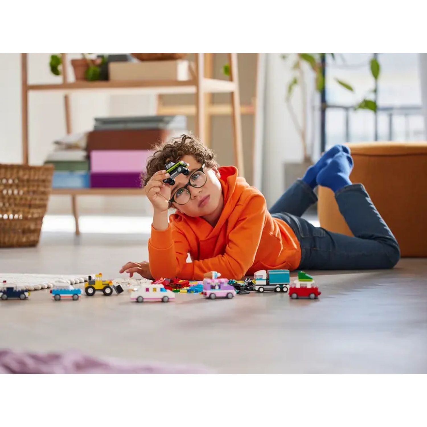 Un garçon en sweat à capuche orange est allongé sur le sol, tenant une voiture jouet. Des voitures jouets sont éparpillées. Une étagère et un panier sont en arrière-plan.