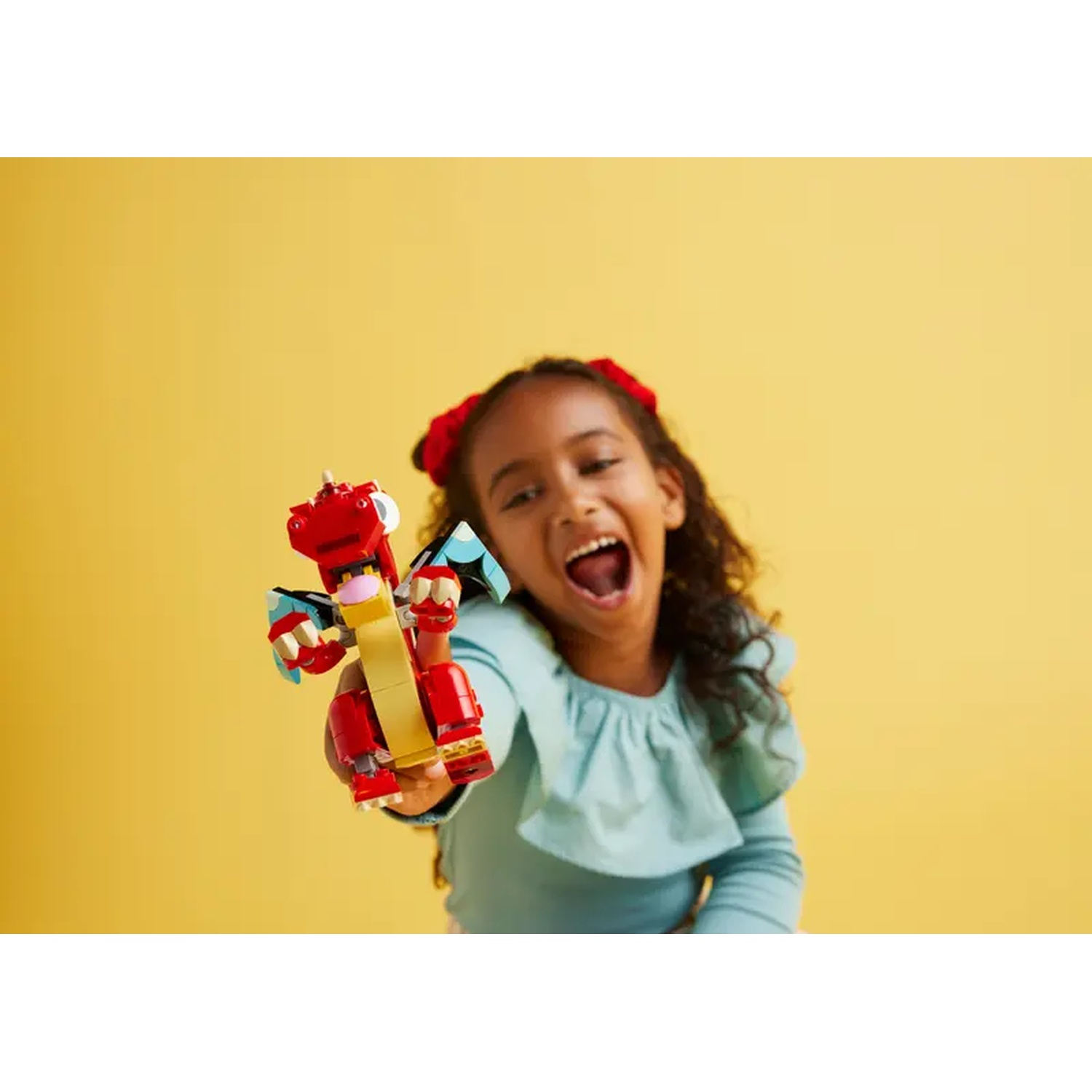 A girl holds up a red Lego dragon toy. She wears a blue shirt and has red bows in her hair, against a yellow background.