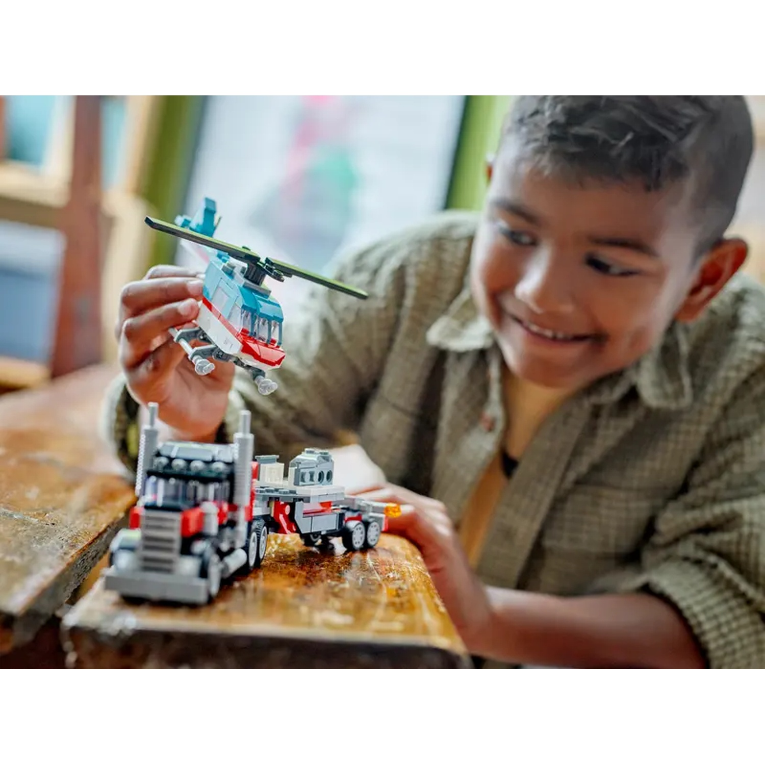 A smiling boy holds a toy helicopter. A toy truck with a trailer is on a wooden surface.