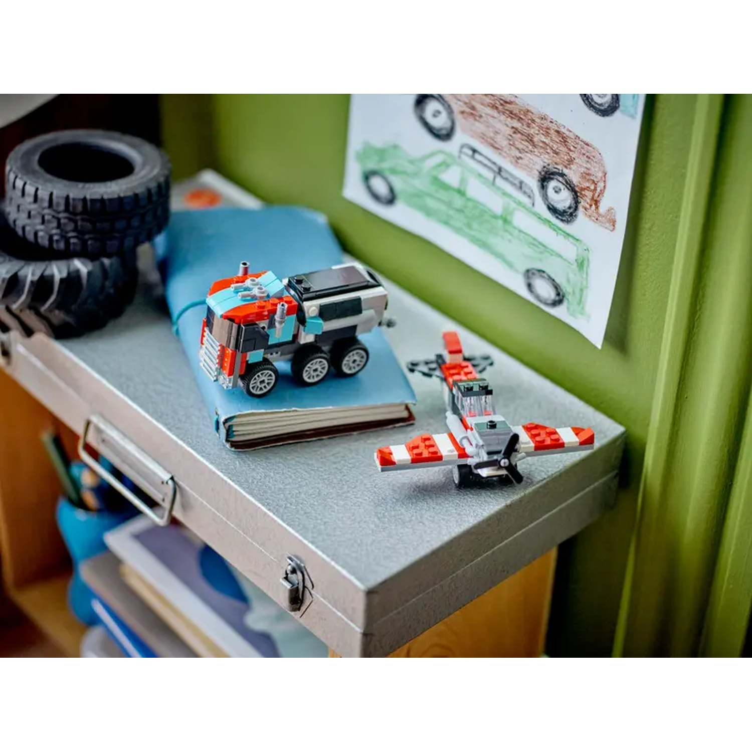 A Lego truck and plane are on a gray surface with tires and a drawing of cars. A blue notebook is visible.