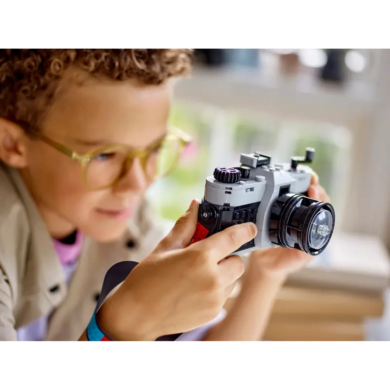 A boy with glasses holds a gray and black toy camera. The camera has a large lens and red accents. He is looking at it.