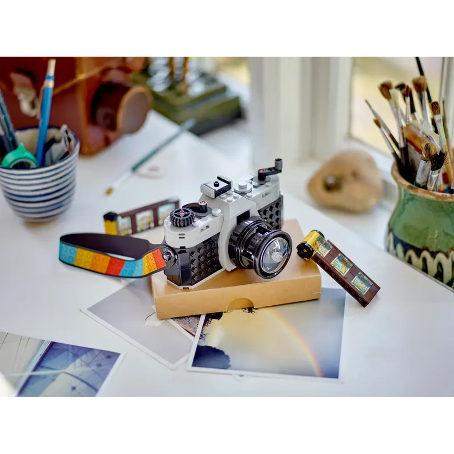 A Lego camera sits on a white table with film strips and photos. A pencil and brushes are in the background.