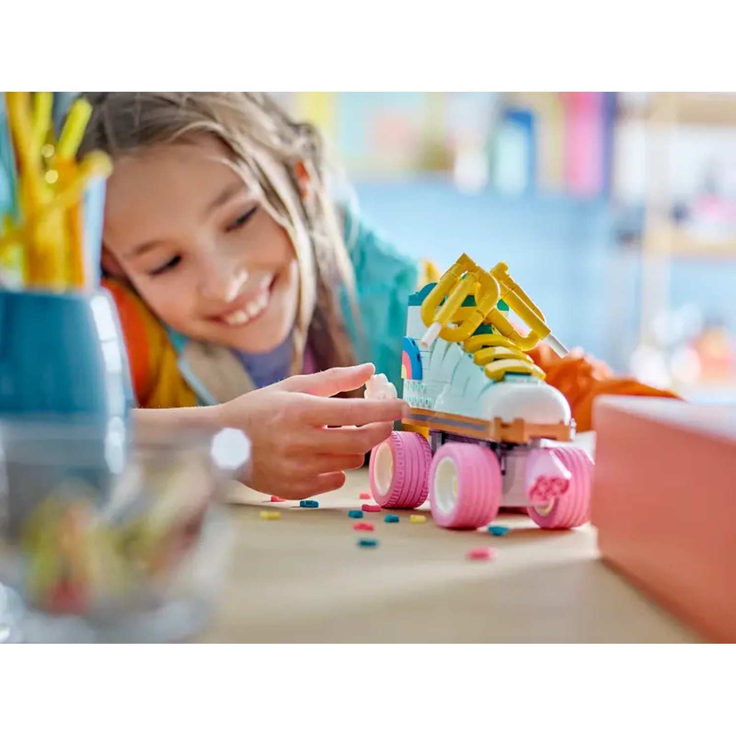 A girl smiles at a toy roller skate made of blocks. The skate is light blue, with pink wheels and yellow laces.