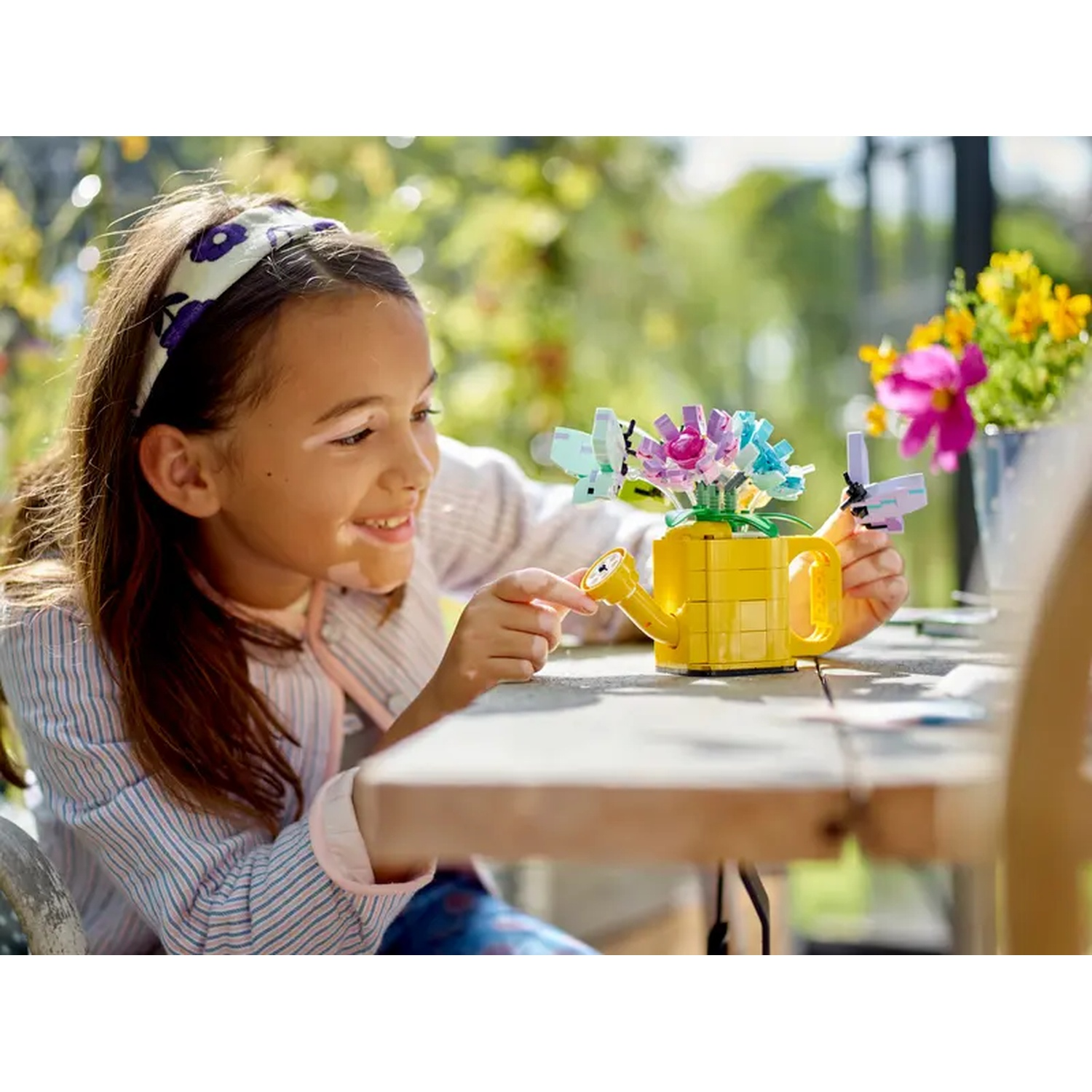 A girl smiles at a Lego yellow watering can with Lego flowers. Other flowers in pots are in the background.