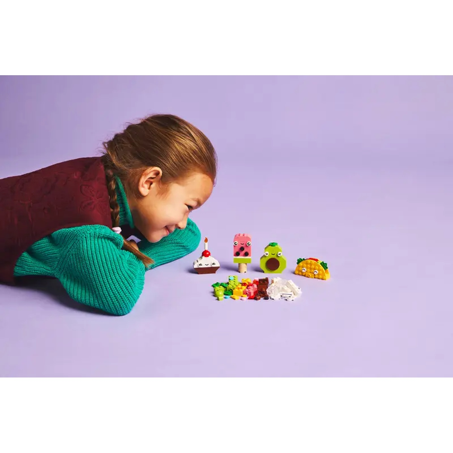 Une fille regarde des aliments Lego : cupcake, glace à la pastèque, avocat et taco. Des blocs Lego colorés sont devant eux, fond violet.