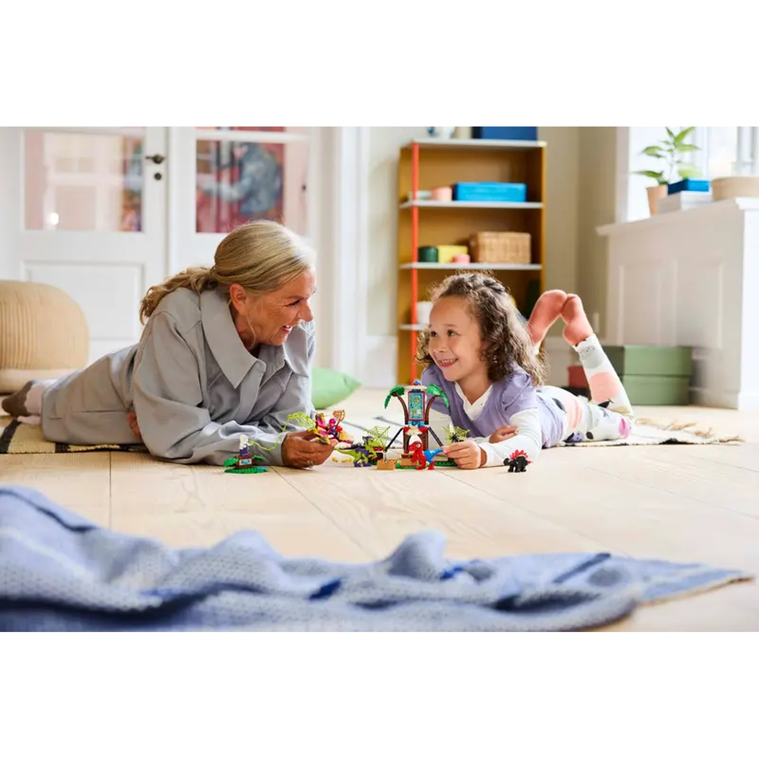 Een vrouw en een meisje spelen met speelgoed op de grond. Ze lachen. Een blauwe deken staat op de voorgrond.