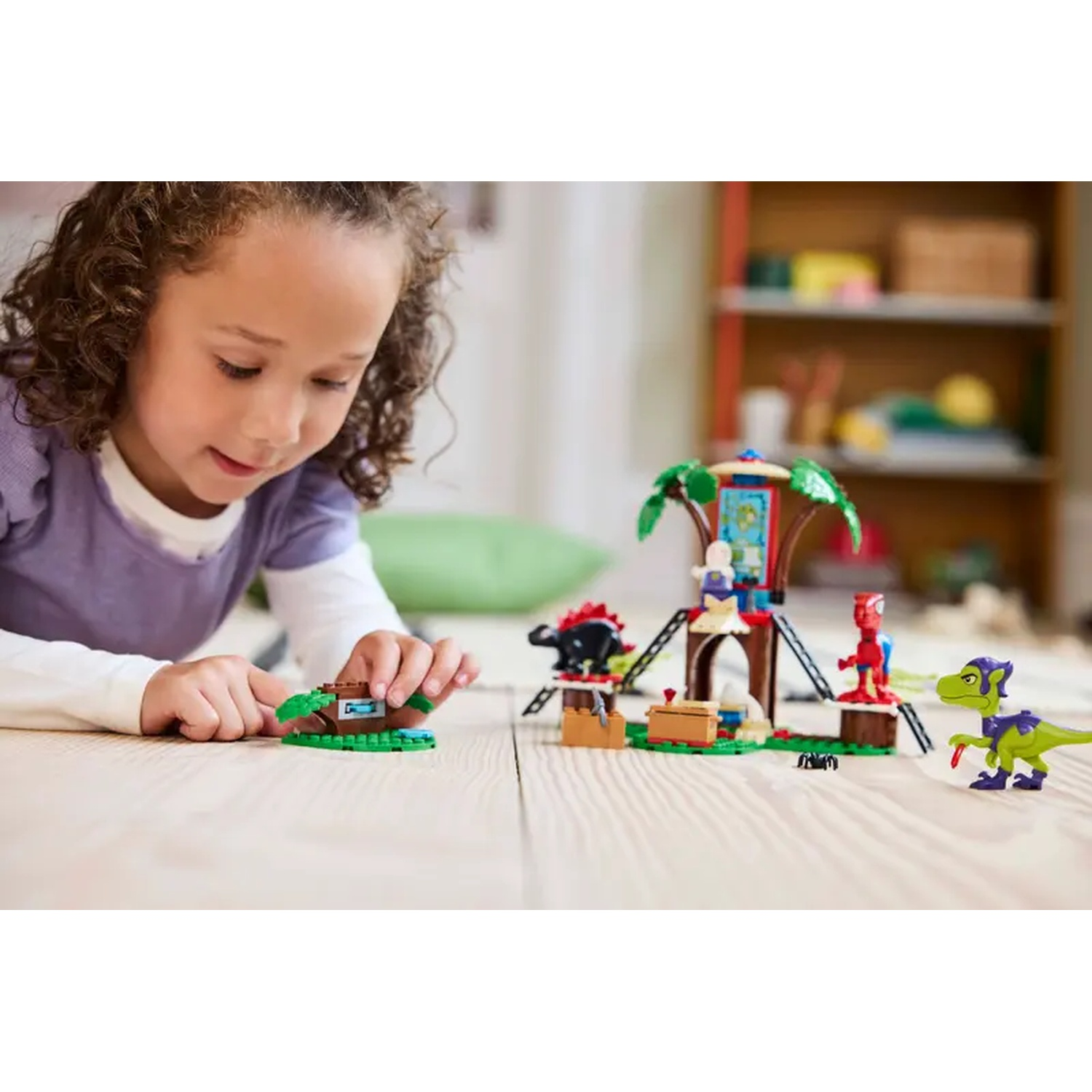 Een jong meisje speelt met een Lego-set, met een boomhut, dinosaurussen en andere figuren. De scène speelt zich af op een houten vloer.