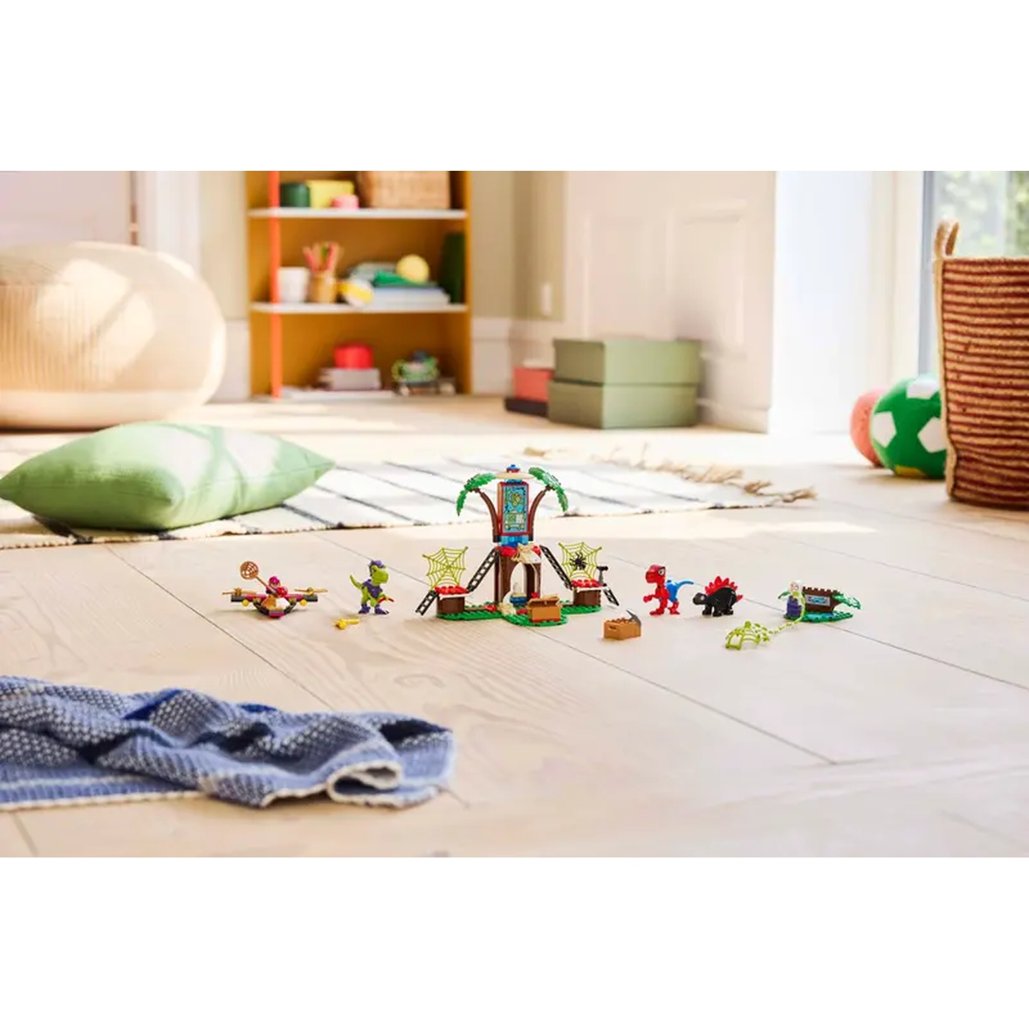 Een Lego dinosaurusset op een houten vloer. Speelgoed omvat een boom, dinosaurussen en een blauwe deken. Ander speelgoed en meubels staan op de achtergrond.