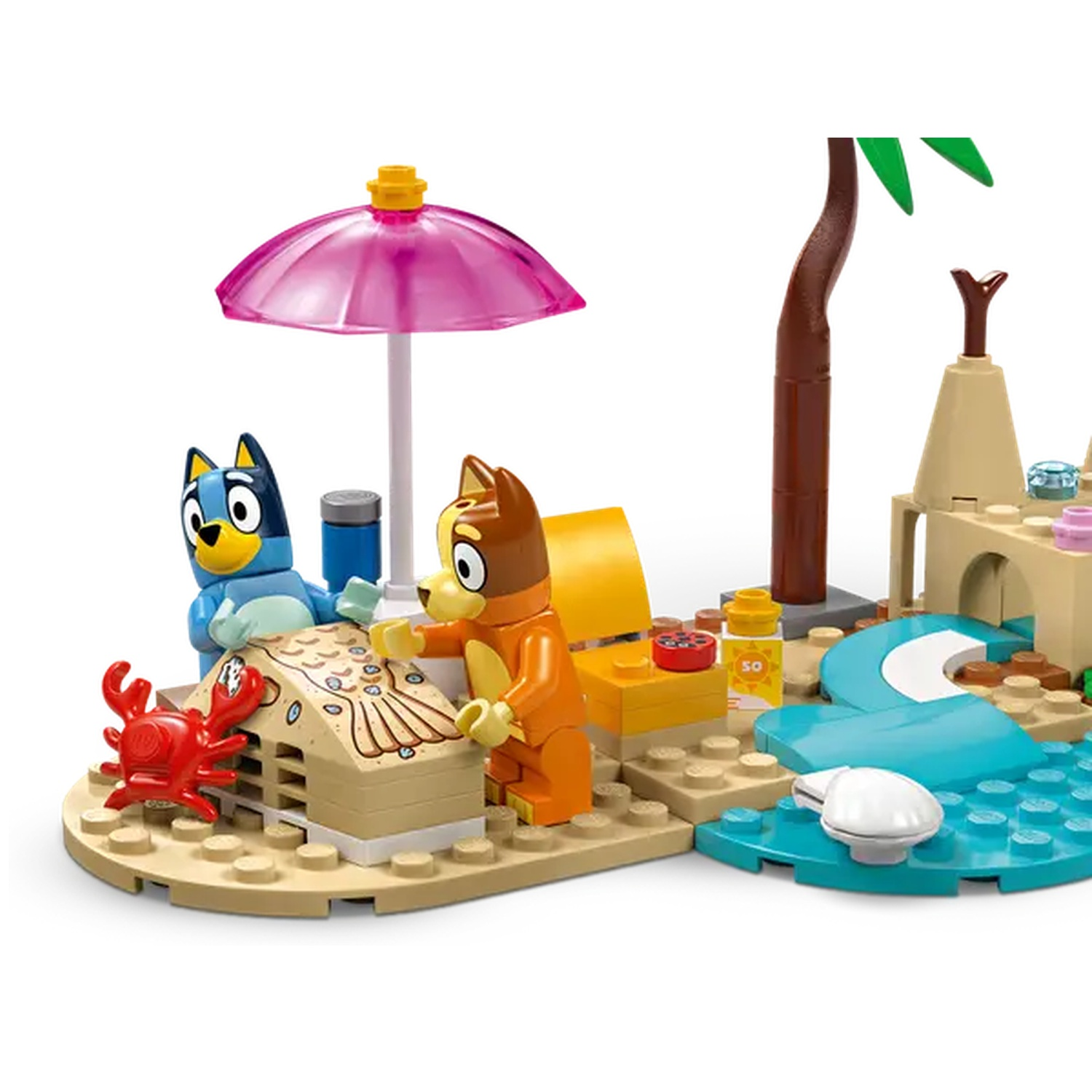 Een Lego strandscène met twee hondenfiguren, een krab, een roze parasol en een palmboom. De honden staan bij een zandkasteel.