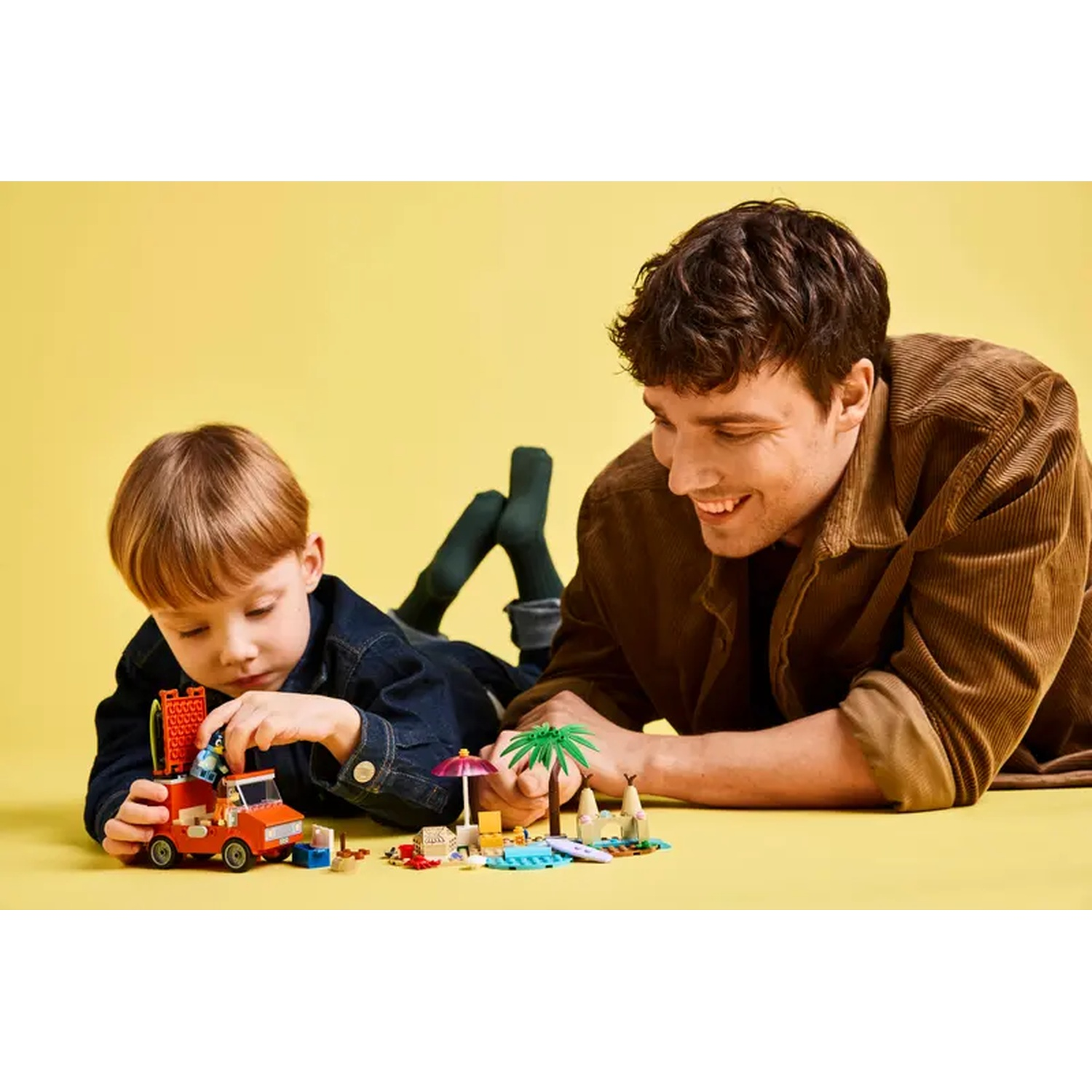 Een man en een jongen spelen met Lego. De jongen zet een auto in elkaar. De man glimlacht naar hem, met een gele achtergrond.