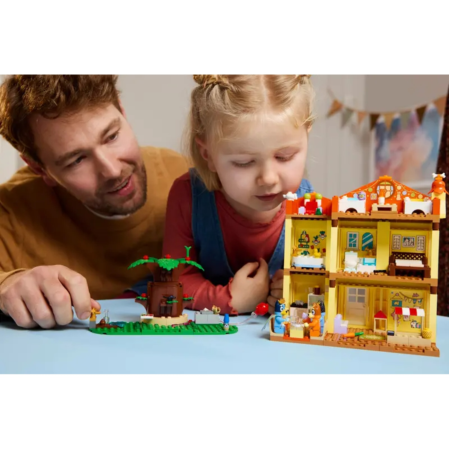 Een man en een meisje spelen met Lego. Ze kijken naar het Lego-huis en een boom. Het huis is geel, met meerdere verdiepingen.