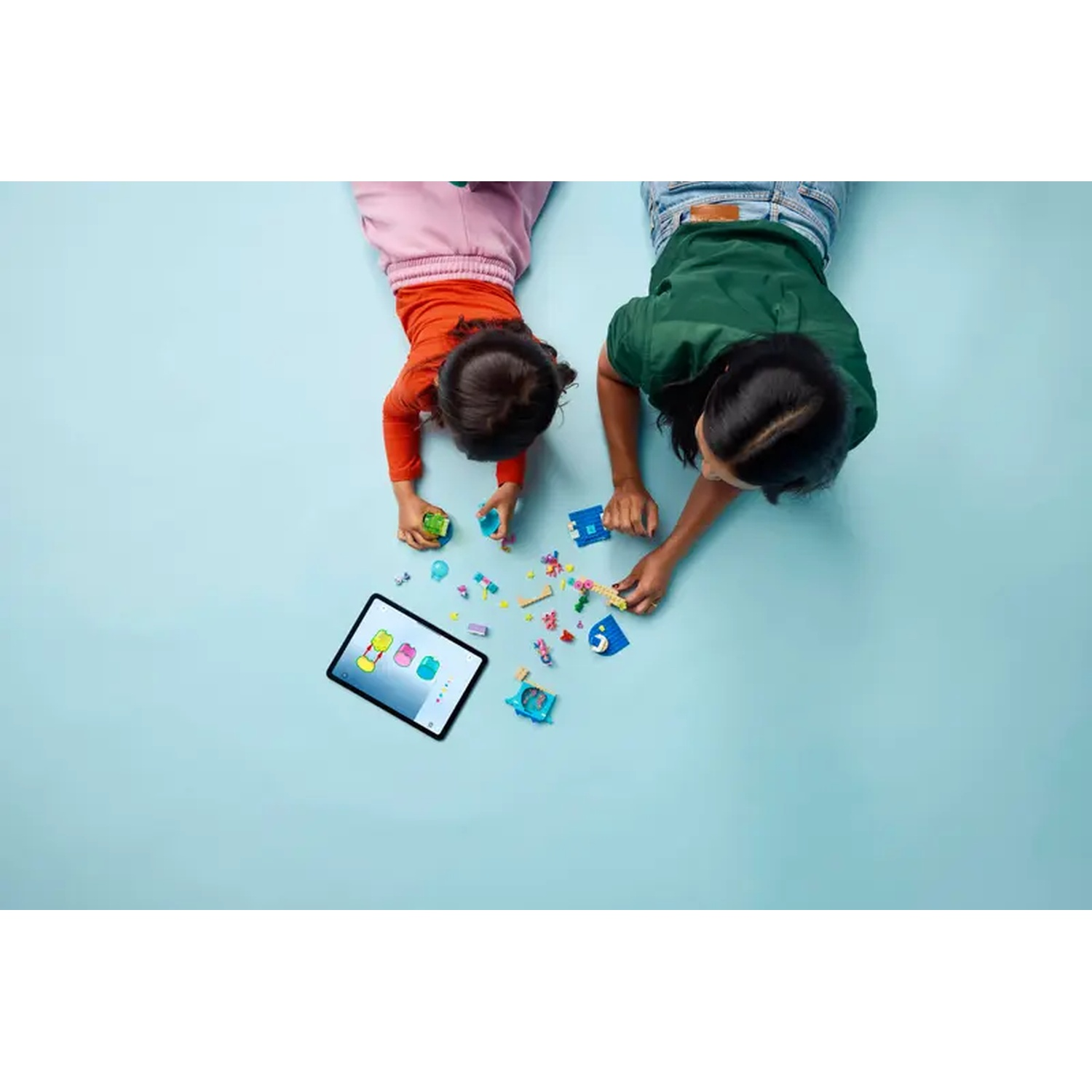 Twee kinderen liggen op een blauw oppervlak en spelen met bouwstenen en een tablet. Eén draagt oranje en roze, de ander groen en blauw.