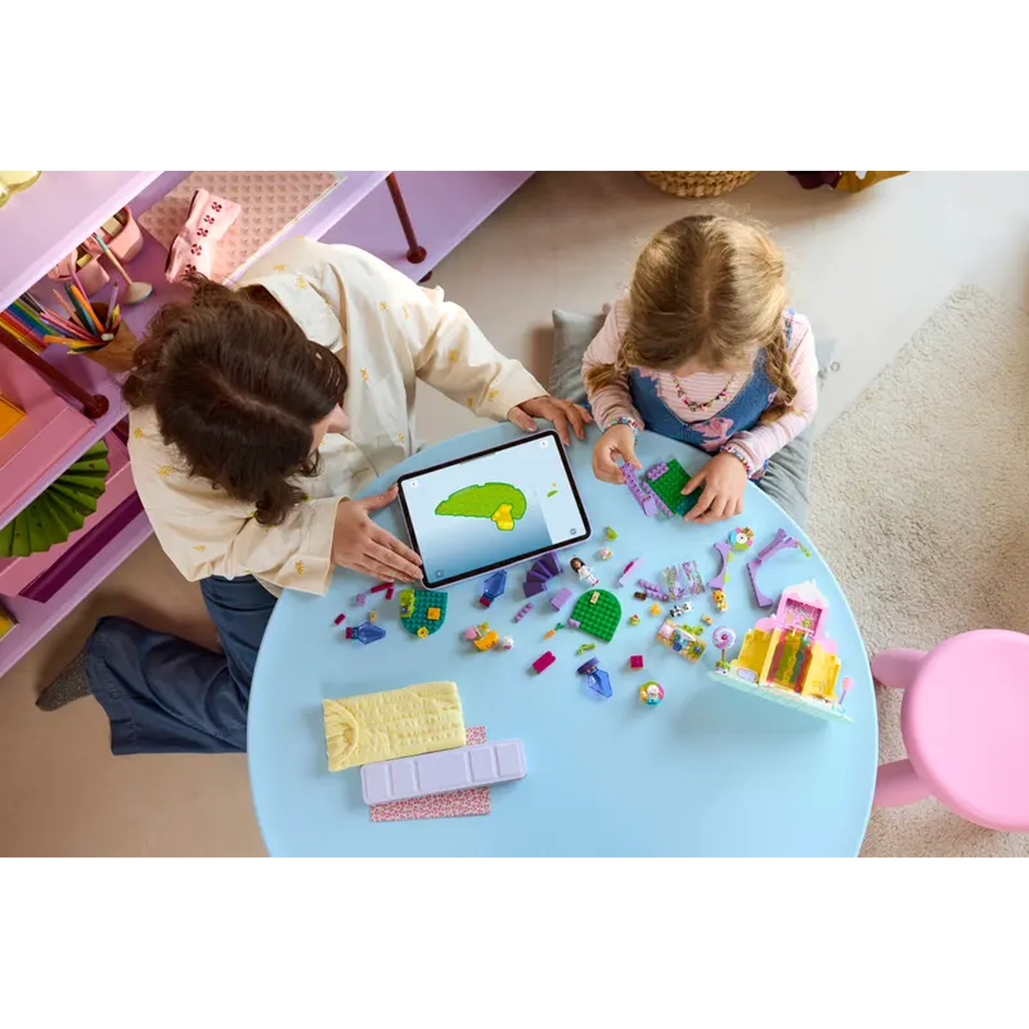 Deux personnes à une table bleue construisant avec des jouets. Une tablette affiche une image verte. Des jouets sont éparpillés sur la table. Chaise rose.