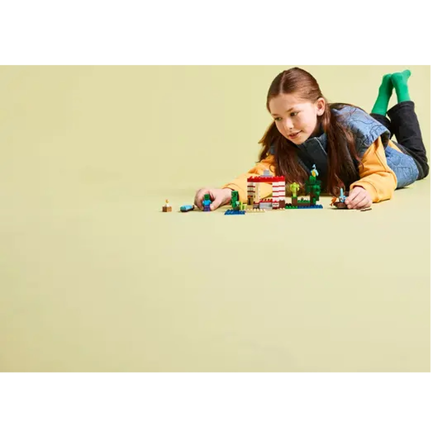 Une fille est allongée sur le ventre en train de jouer avec des LEGO. Elle porte une chemise jaune, un gilet en jean et un pantalon noir. Des chaussettes vertes sont visibles.