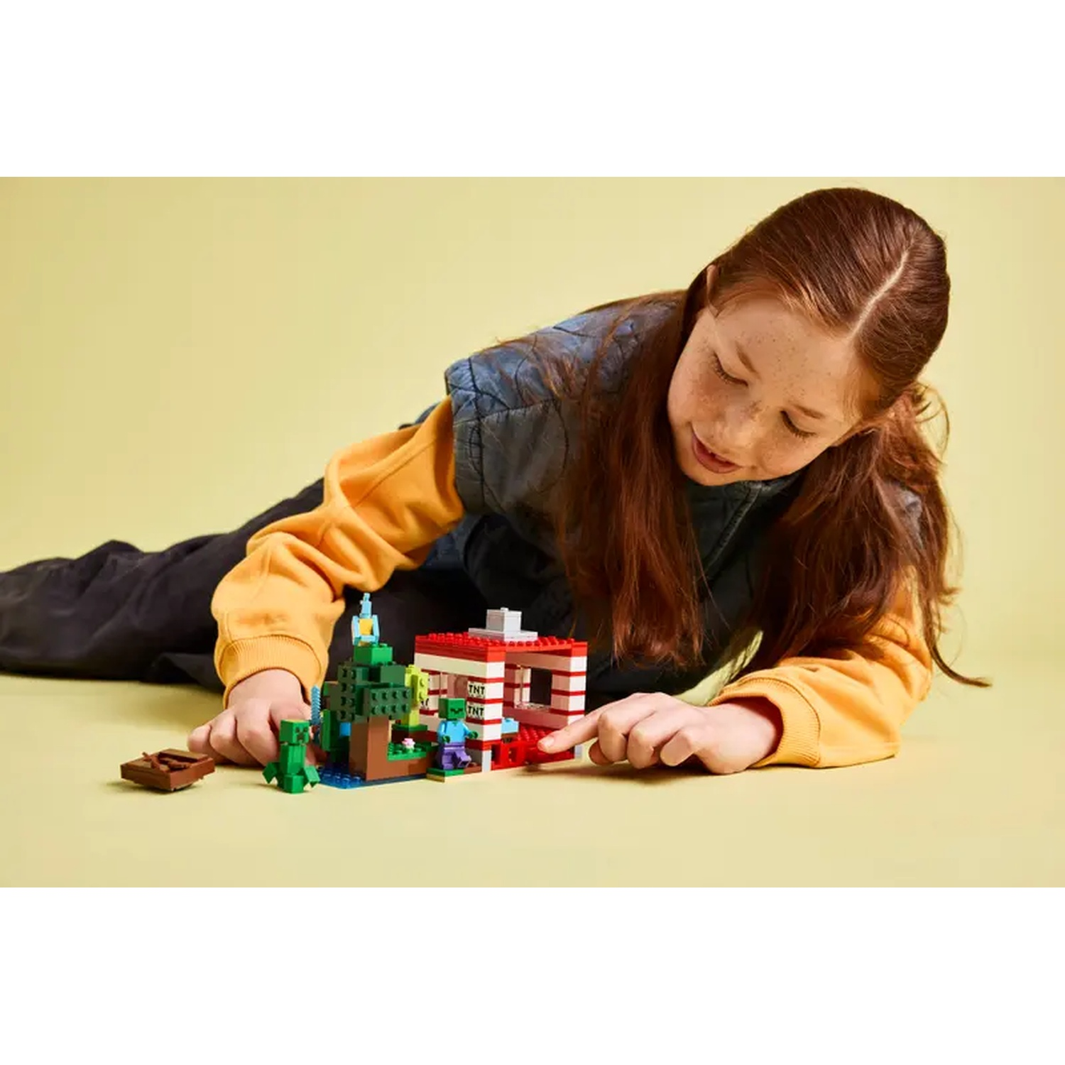 Une fille joue avec un set Lego Minecraft au sol. L'ensemble comprend une maison, des arbres, des personnages et un bateau.