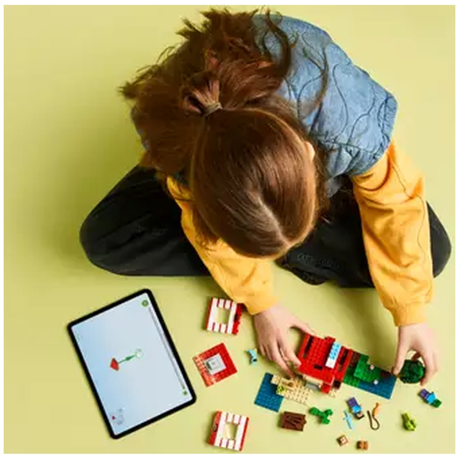 Une personne construit avec des briques Lego, une tablette affichant des instructions est à proximité, sur une surface vert clair.