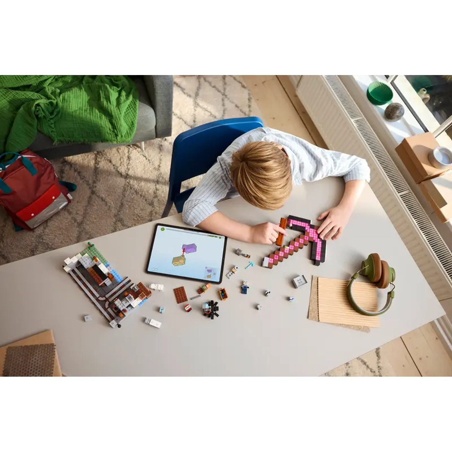 Un enfant construit avec des blocs Minecraft, avec une tablette à proximité. Une pioche, un casque et d'autres blocs sont sur une table grise.