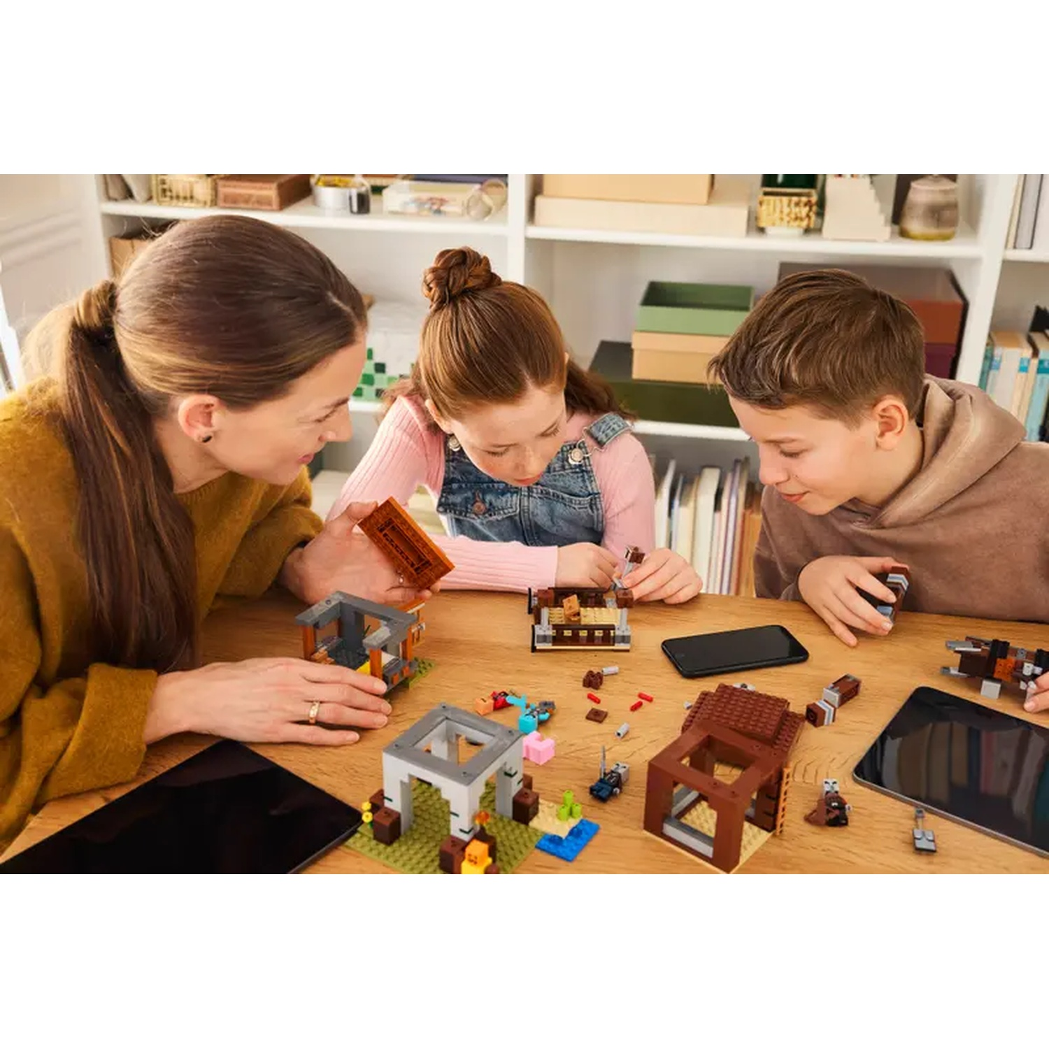 Trois personnes construisant avec des Lego sur une table en bois. Il y a des structures Lego, une tablette et un smartphone. Une étagère est en arrière-plan.