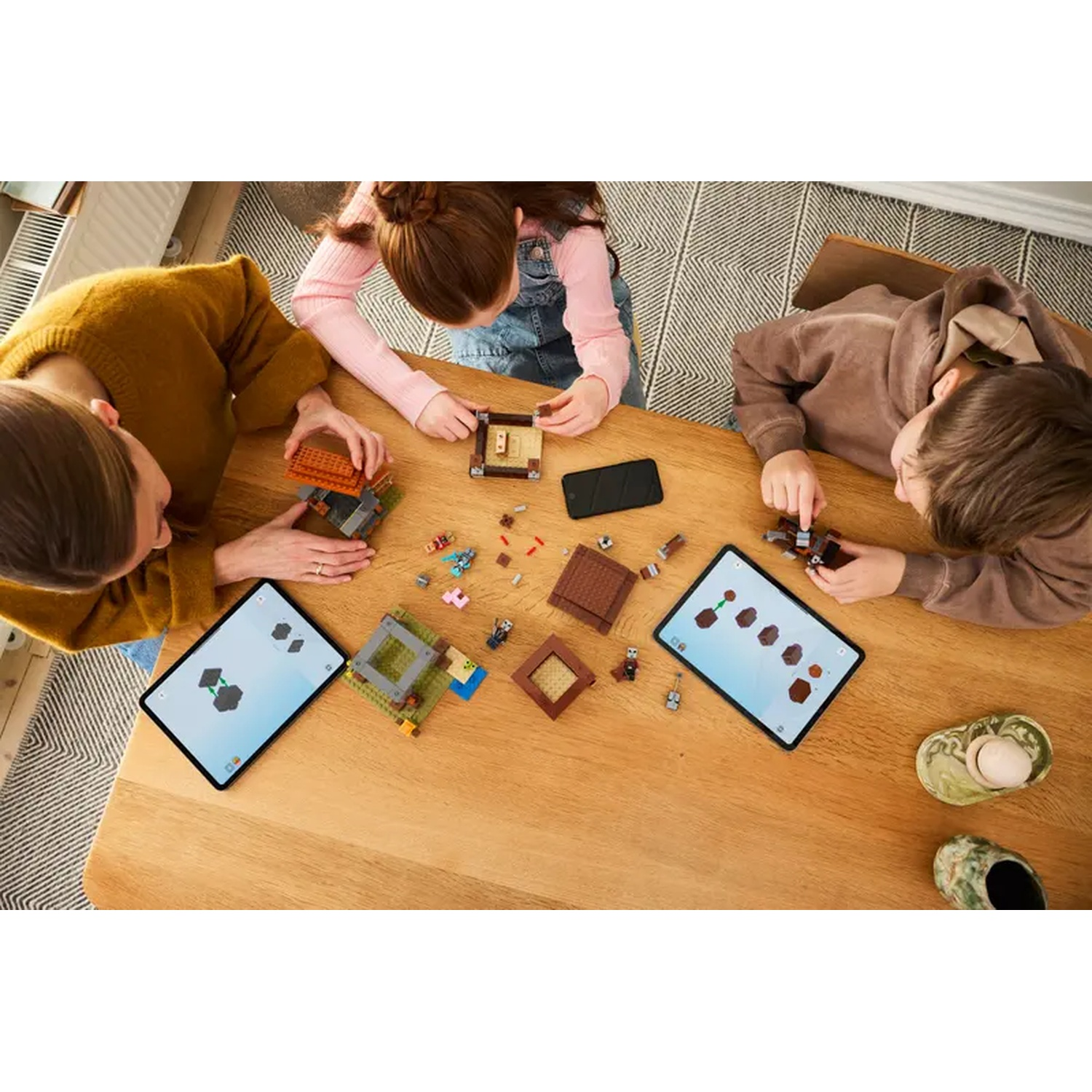 Trois personnes construisent avec Lego sur une table en bois. Il y a deux tablettes, un téléphone et des pièces Lego éparpillées.