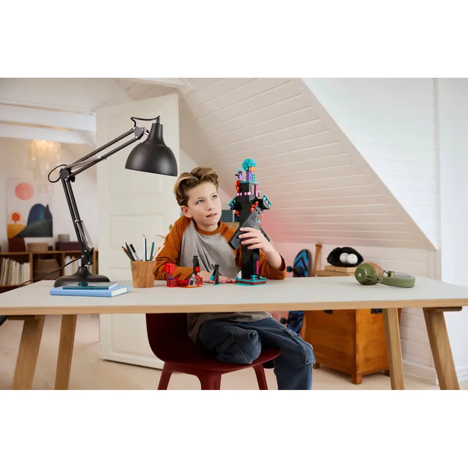 Un garçon est assis à un bureau, construisant avec des blocs colorés. Une lampe, des crayons, des livres et un casque sont à proximité.