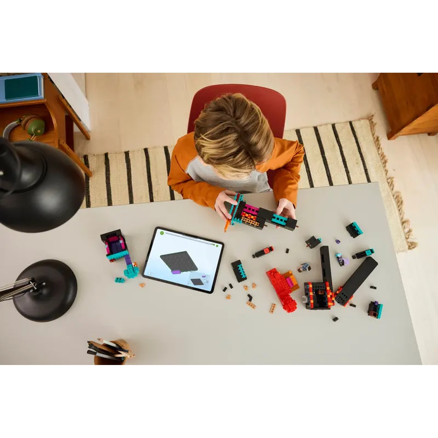 Un enfant construit avec des blocs colorés sur une table. Une tablette affichant un guide de construction est à proximité. Une lampe et un tapis sont également présents.