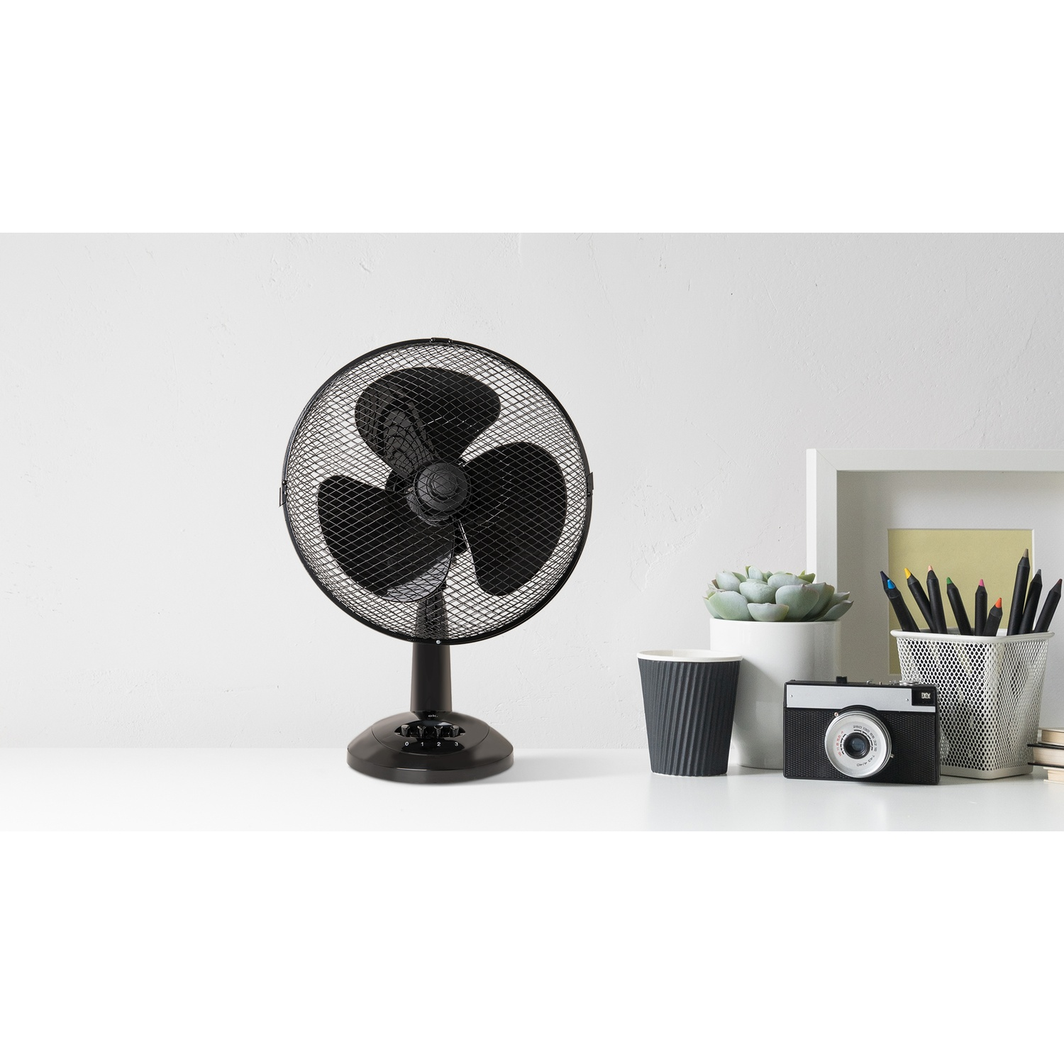 Un ventilateur de bureau noir, un appareil photo, une plante succulente, une tasse et des crayons sont sur un bureau blanc contre un mur blanc.