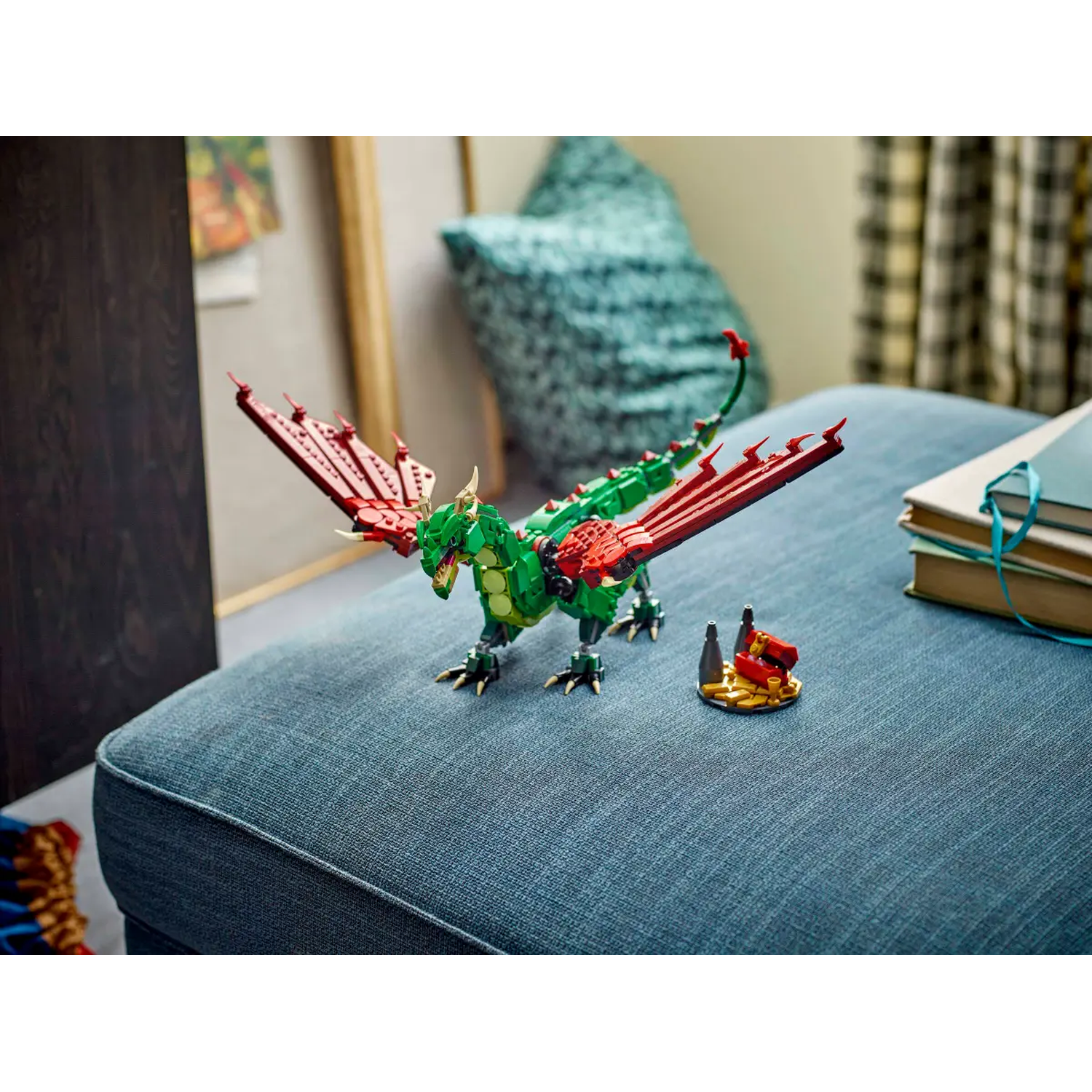 A Lego dragon with red wings and a green body sits on a blue ottoman. A small Lego structure and books are in the background.
