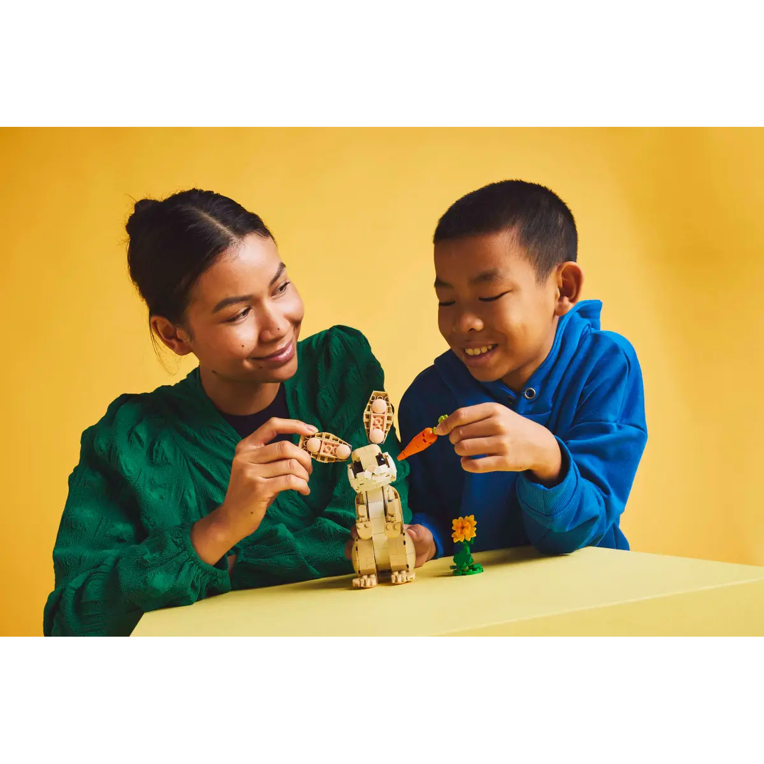 A woman and a boy build a Lego bunny. The bunny, carrot, and flower are on a yellow table. Yellow background.