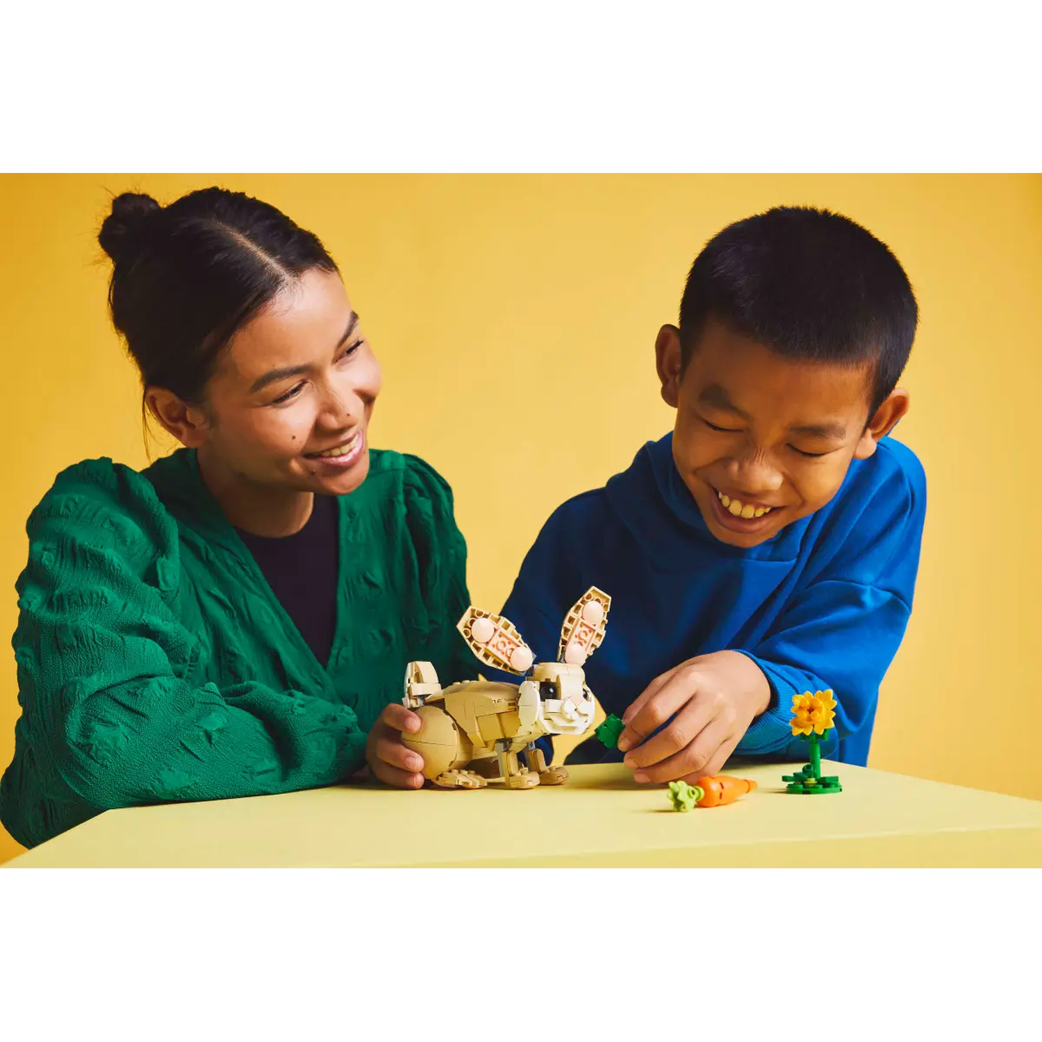 A woman and a boy smile at a Lego rabbit. The boy holds a carrot for the rabbit. A Lego flower and carrot are on the table.