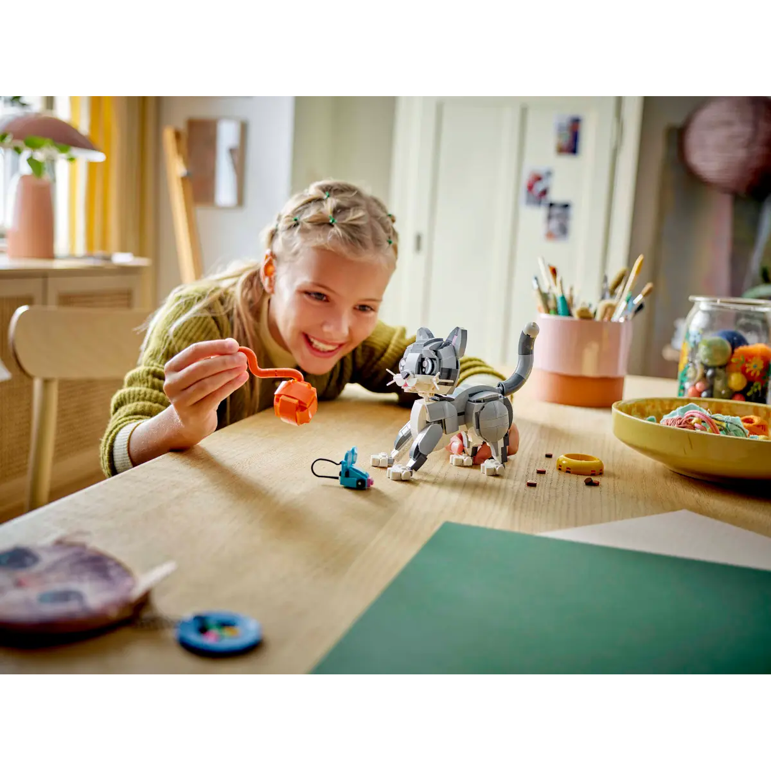 Een meisje lacht naar een LEGO-kat. De kat staat op een tafel met speelgoed en kunstbenodigdheden. Het meisje houdt een oranje LEGO-stuk vast.