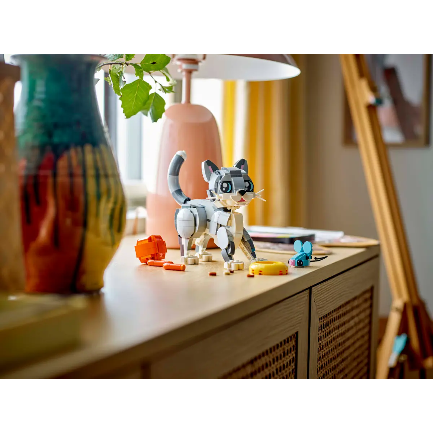 Een Lego-kat, oranje blokken en een blauwe muis staan op een houten oppervlak. Een vaas en een lamp staan op de achtergrond.