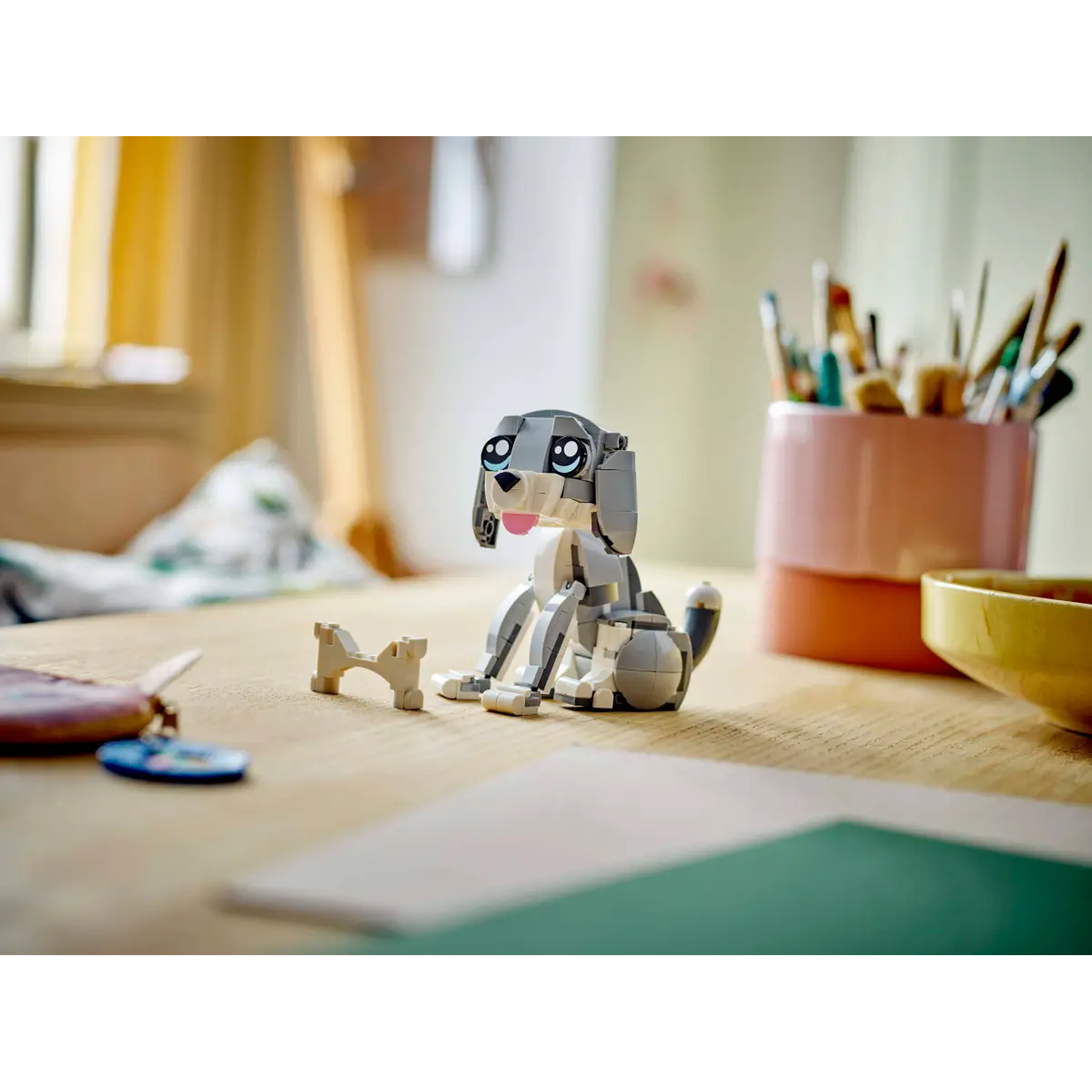Een Lego-hond zit op een tafel met een bot, penselen en een gele kom op de achtergrond. De hond is grijs en wit.