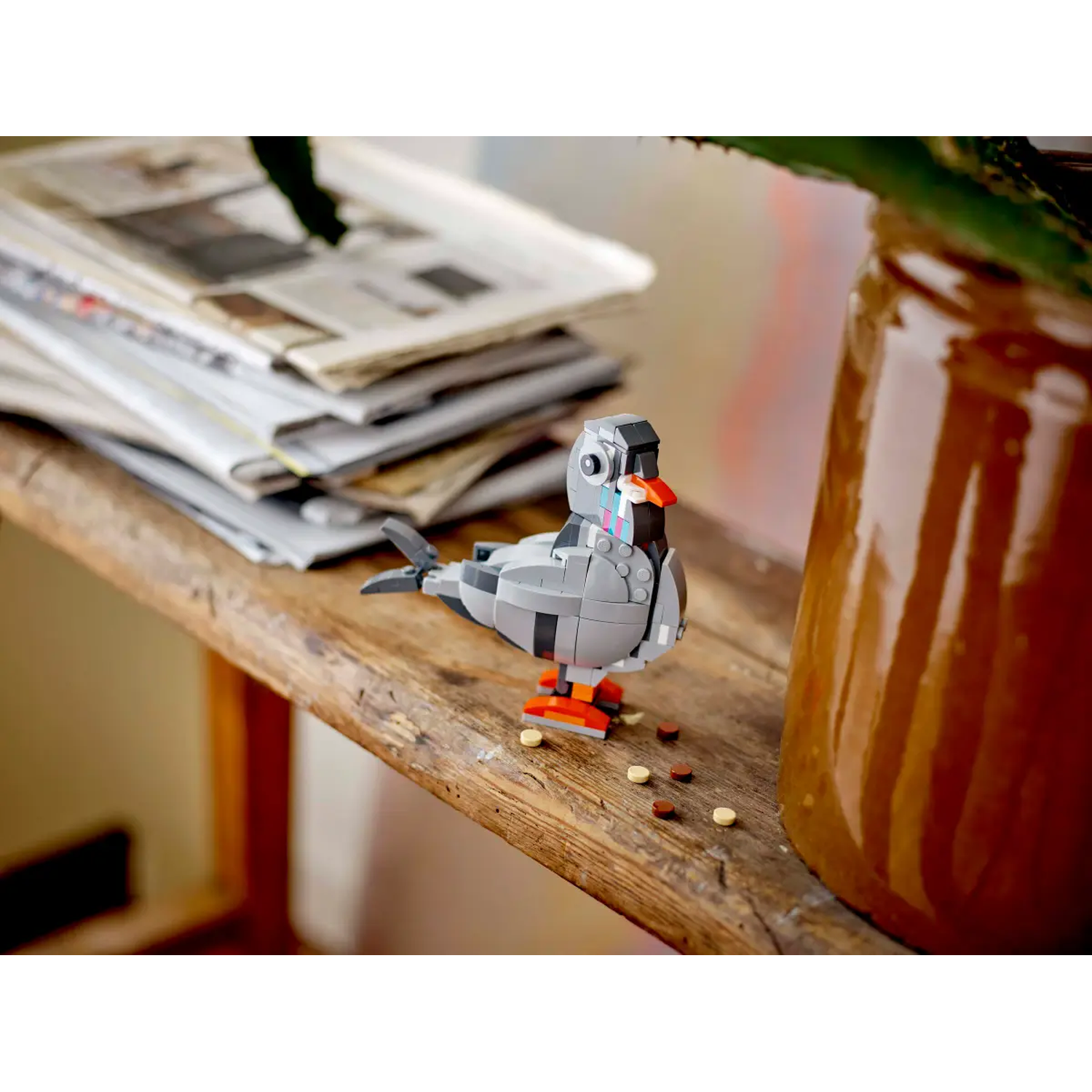 Een Lego-duif zit op een houten plank, naast een bruine pot en kranten. De duif is grijs met oranje poten en een rode snavel.