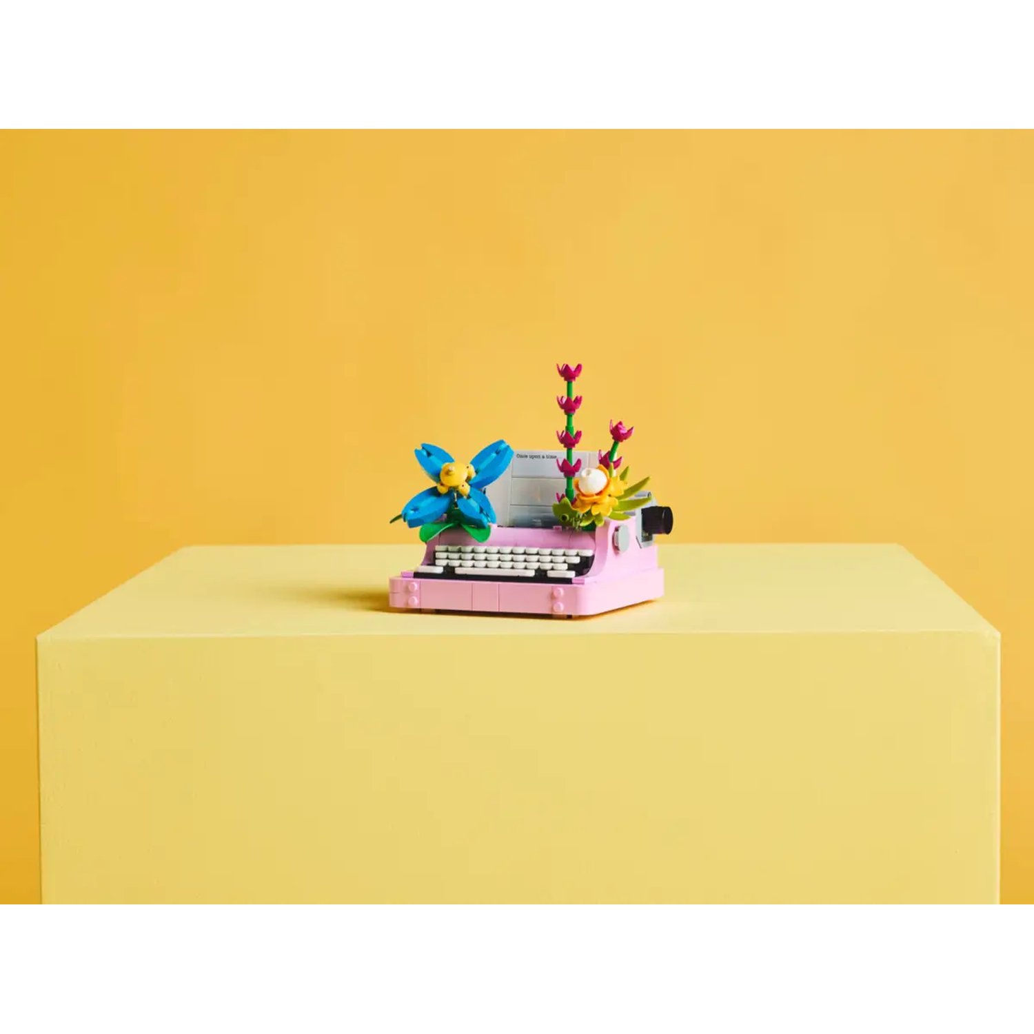 A pink Lego typewriter with blue flower and other Lego flowers. It sits on a yellow block, against a yellow background.