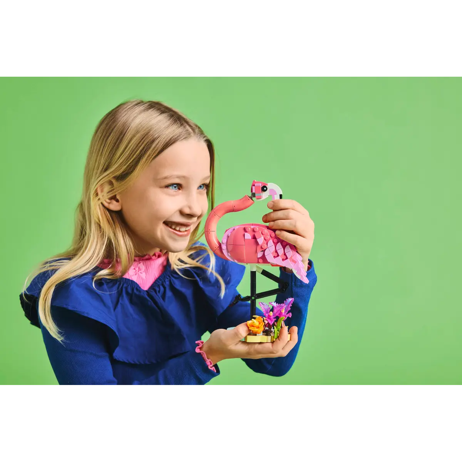 A girl smiles, holding a Lego flamingo and flowers against a green background. She wears a blue top with pink trim.