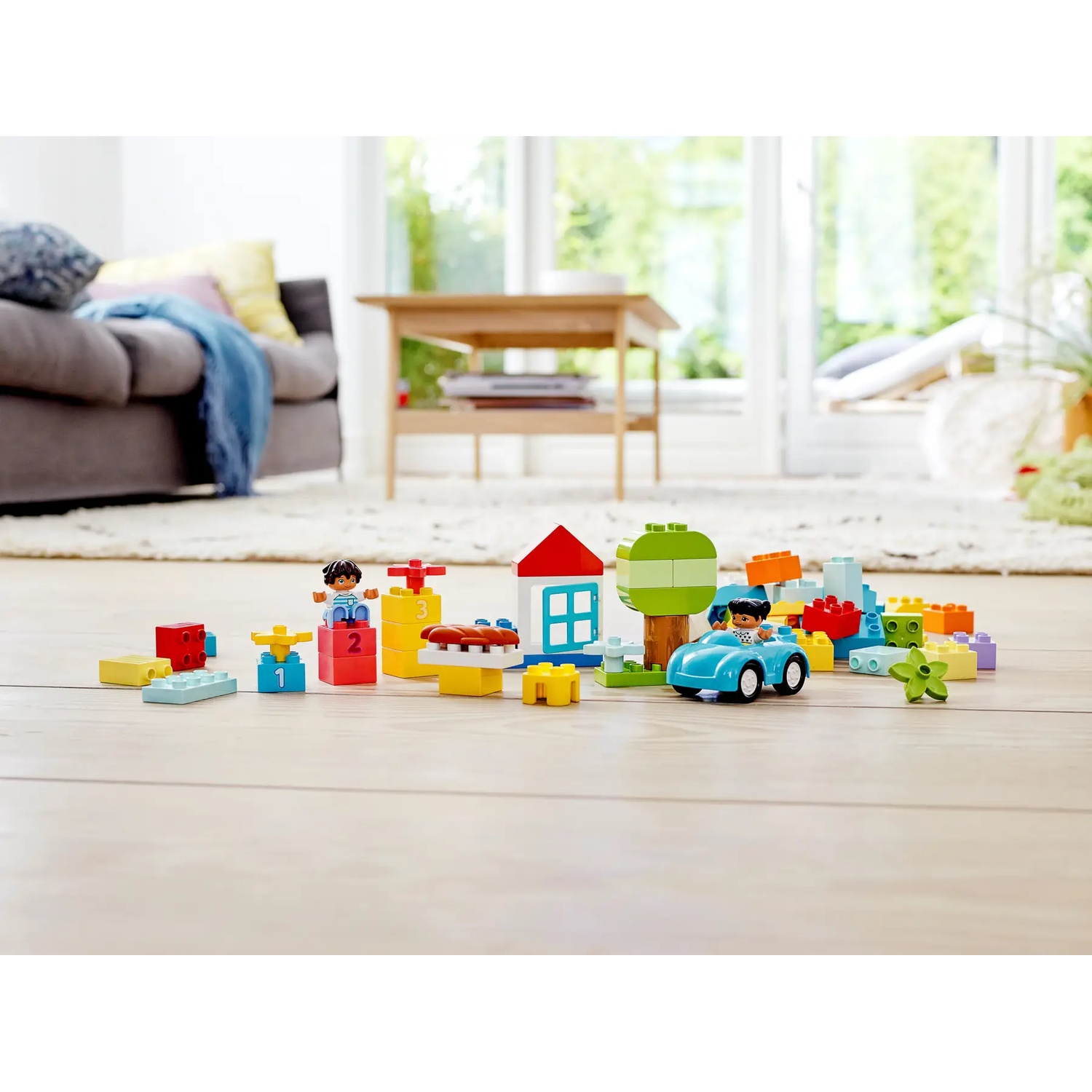 Blocs de jouets colorés disposés sur un sol en bois. Comprend une voiture bleue avec une figurine, une maison, un arbre et d'autres blocs.