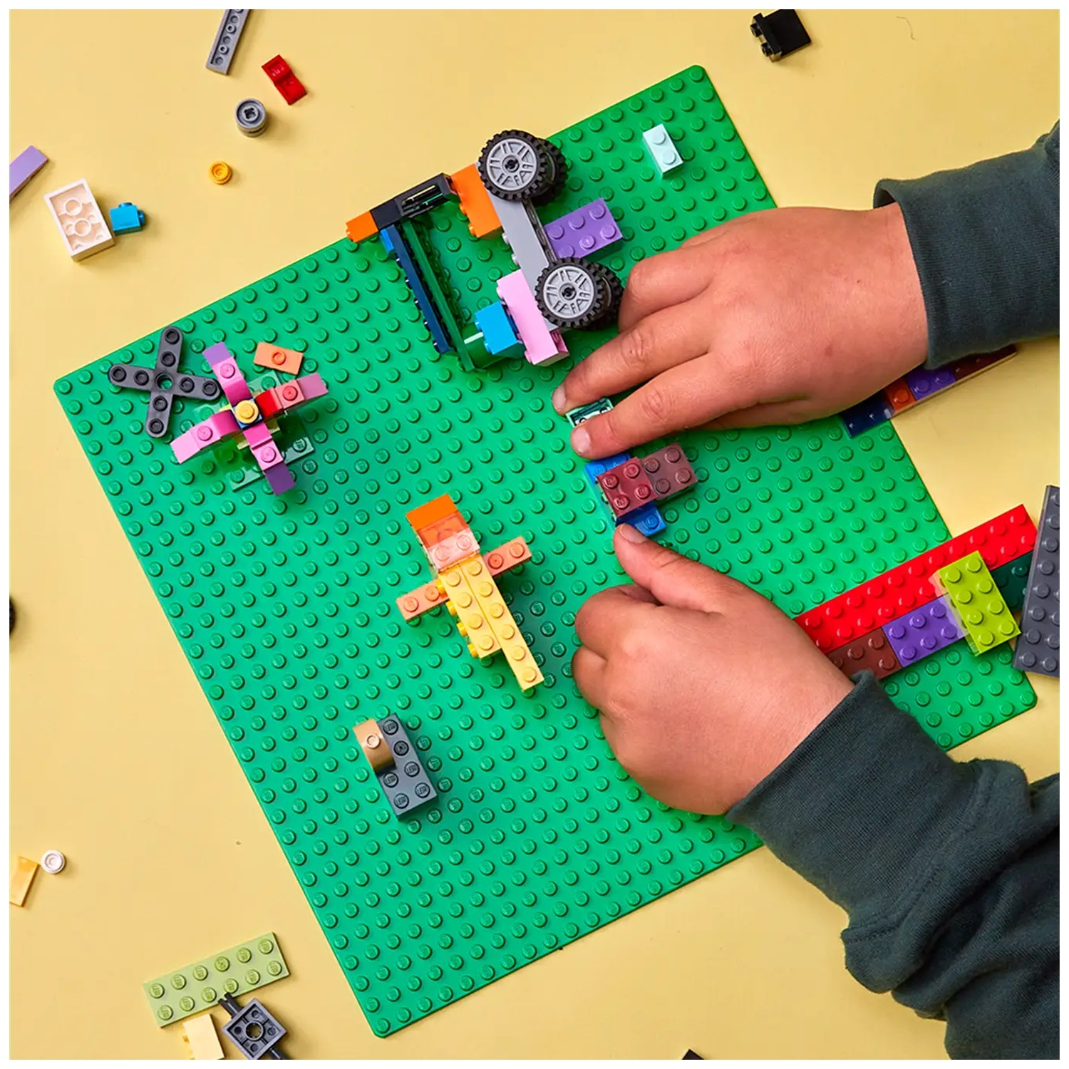 Mains construisant avec des briques Lego colorées sur une plaque verte, fond jaune. Diverses pièces Lego éparpillées.