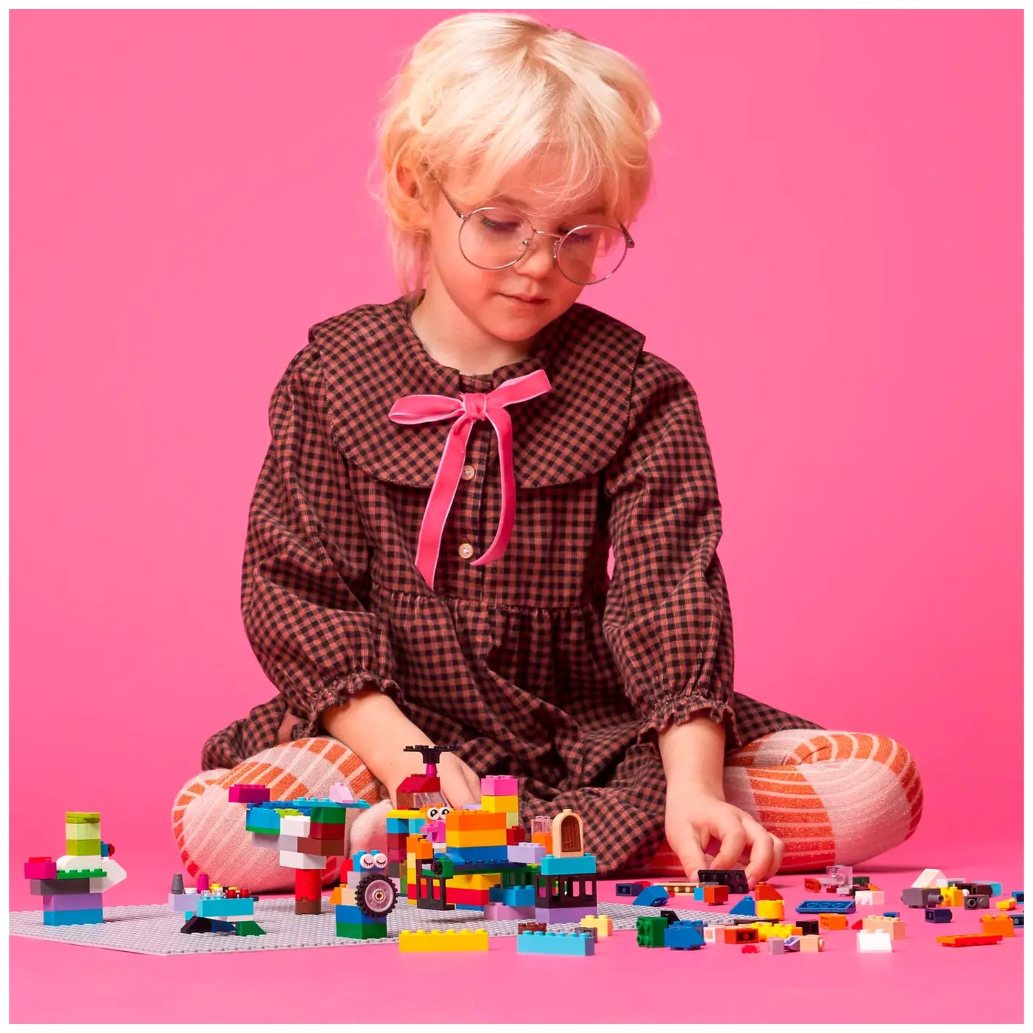 Een kind met een bril en blond haar zit omringd door kleurrijke speelgoedblokken op een roze achtergrond, gefocust op bouwen.