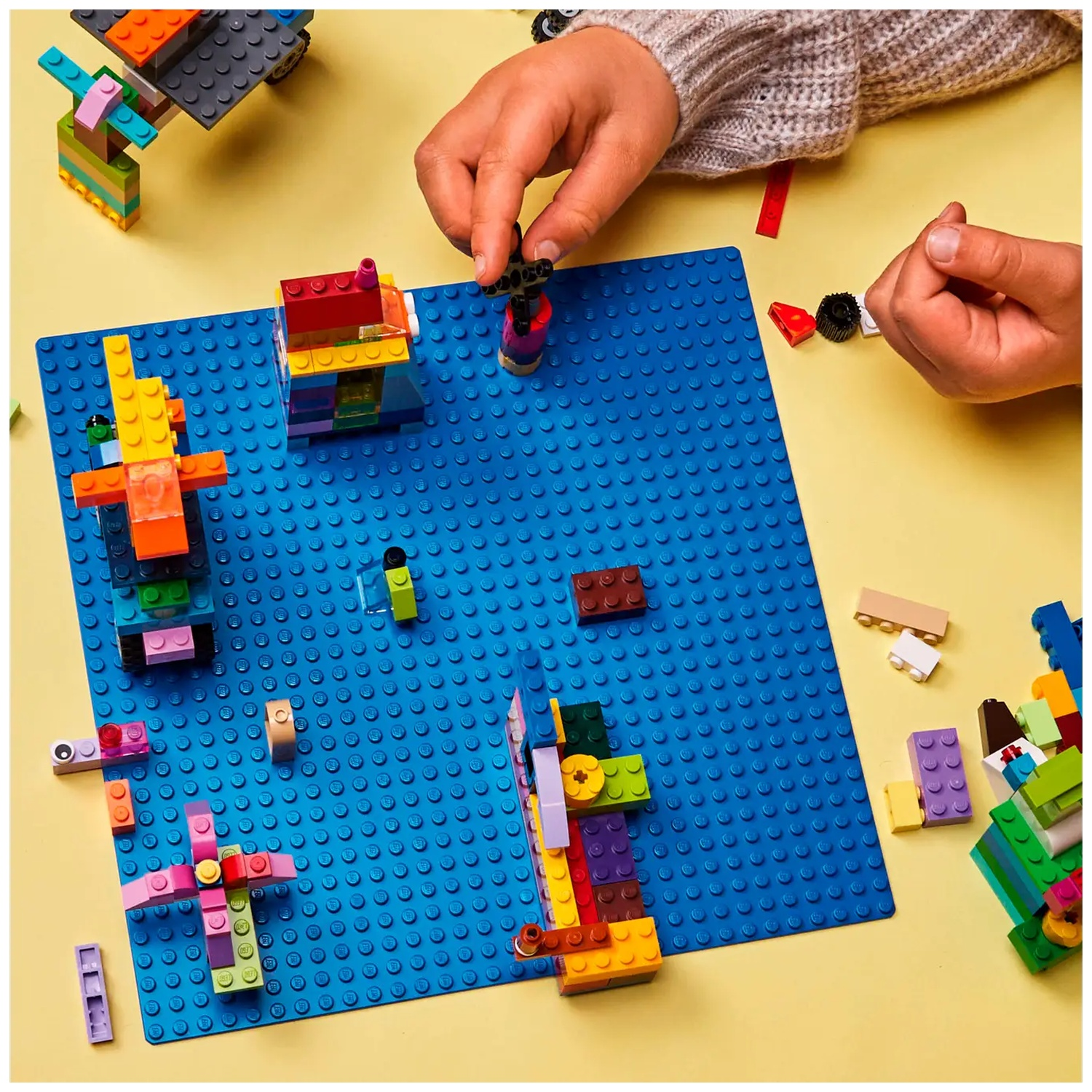 Mains construisant avec des blocs colorés sur une plaque de base bleue. Diverses structures sont partiellement construites. Fond jaune.