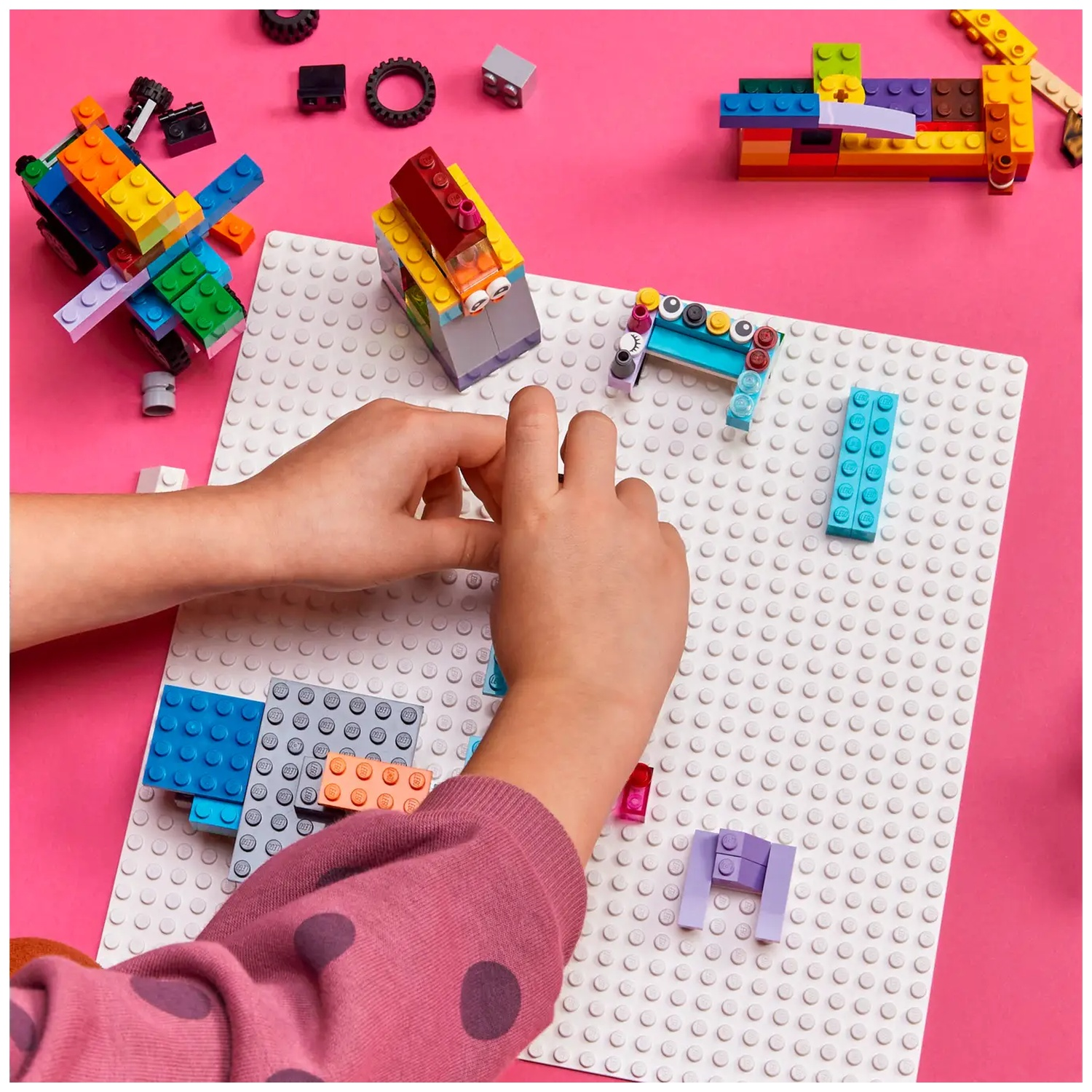 Un enfant construit avec des briques Lego colorées sur une base blanche. Fond rose. Diverses structures Lego sont visibles.