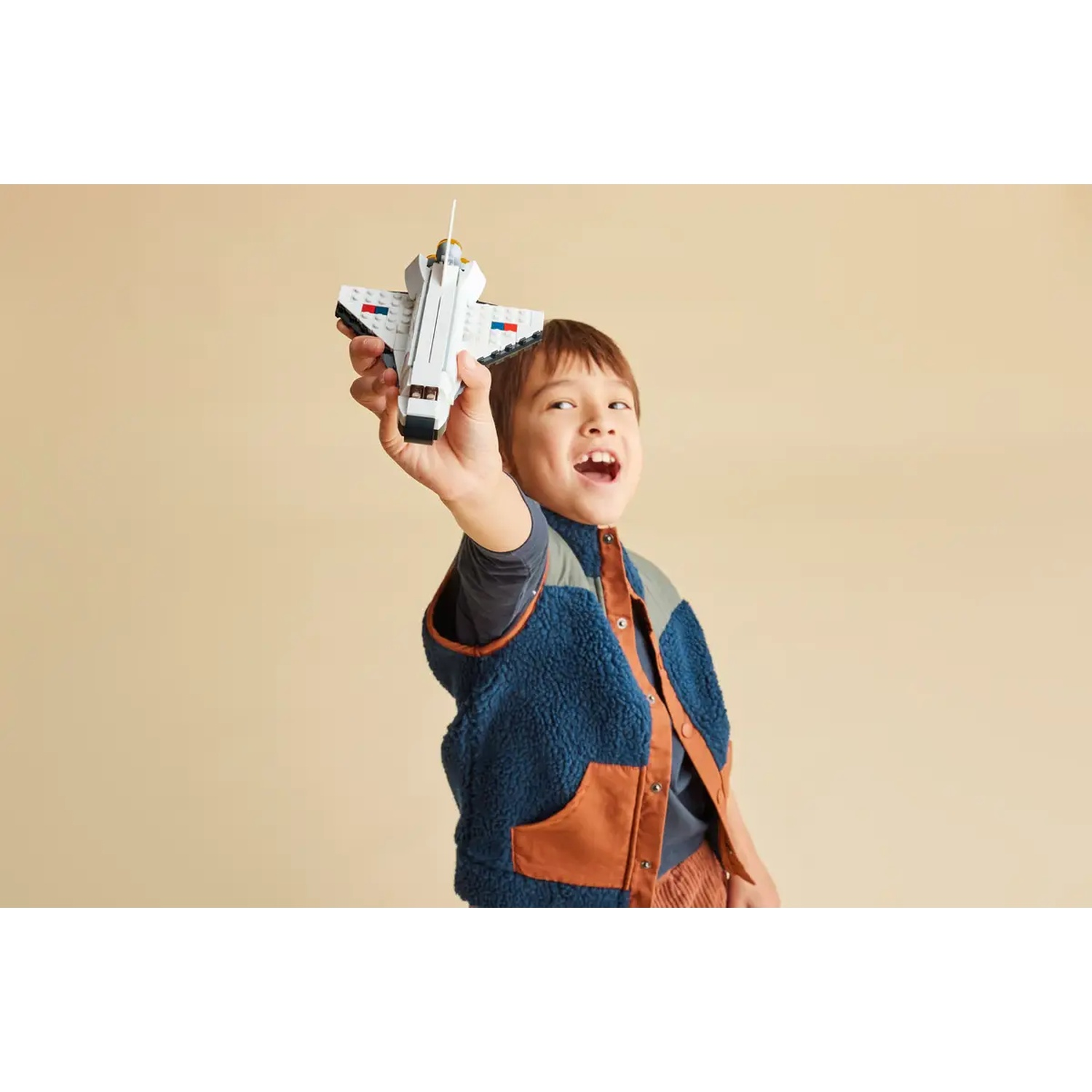 A boy holds up a toy space shuttle. He wears a blue vest, brown corduroy pants, and a gray shirt. The background is tan.