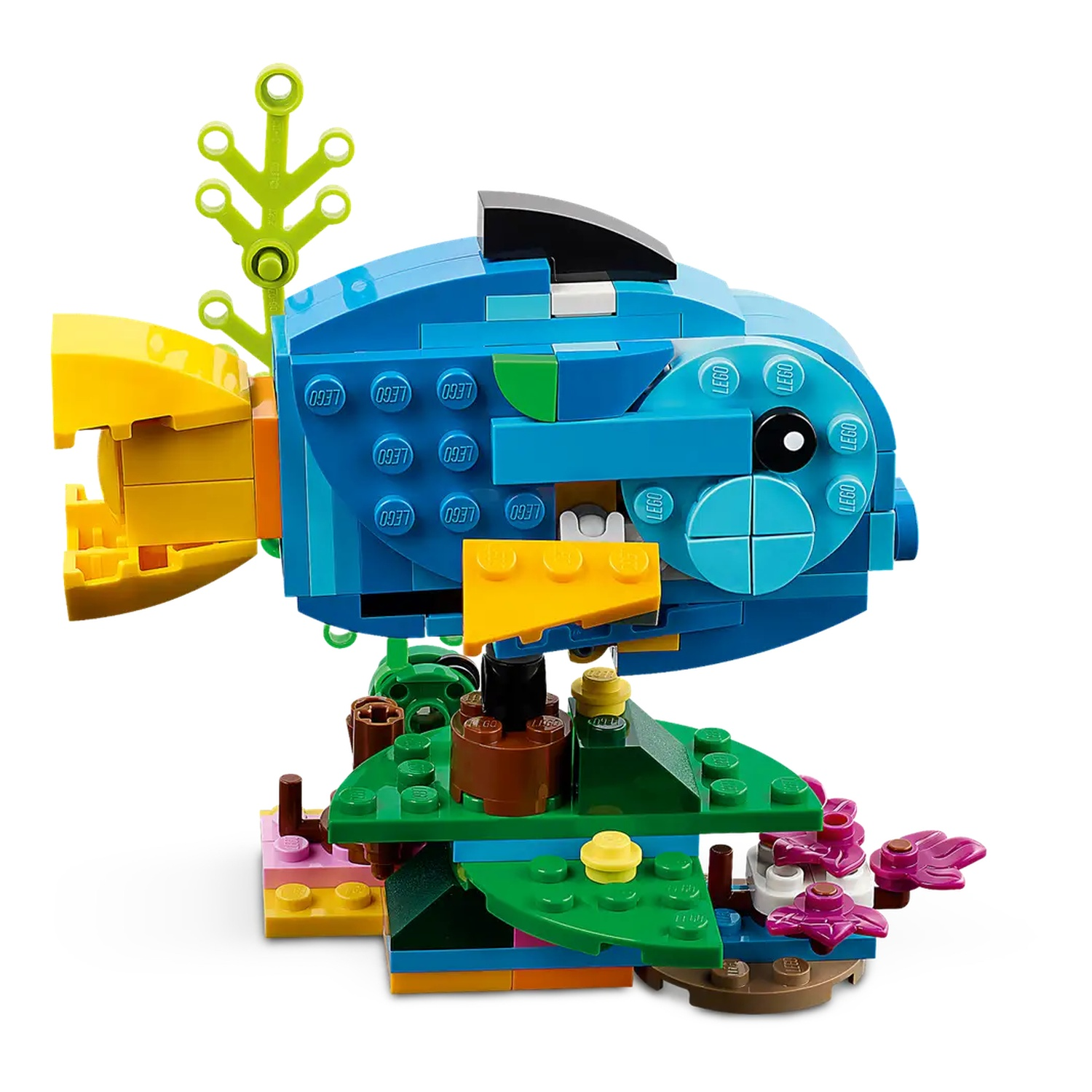 Een Lego vis, blauw lichaam, gele staart en zwarte vin. Het staat op een groene basis met bruine en roze blokken, plus bloemen.