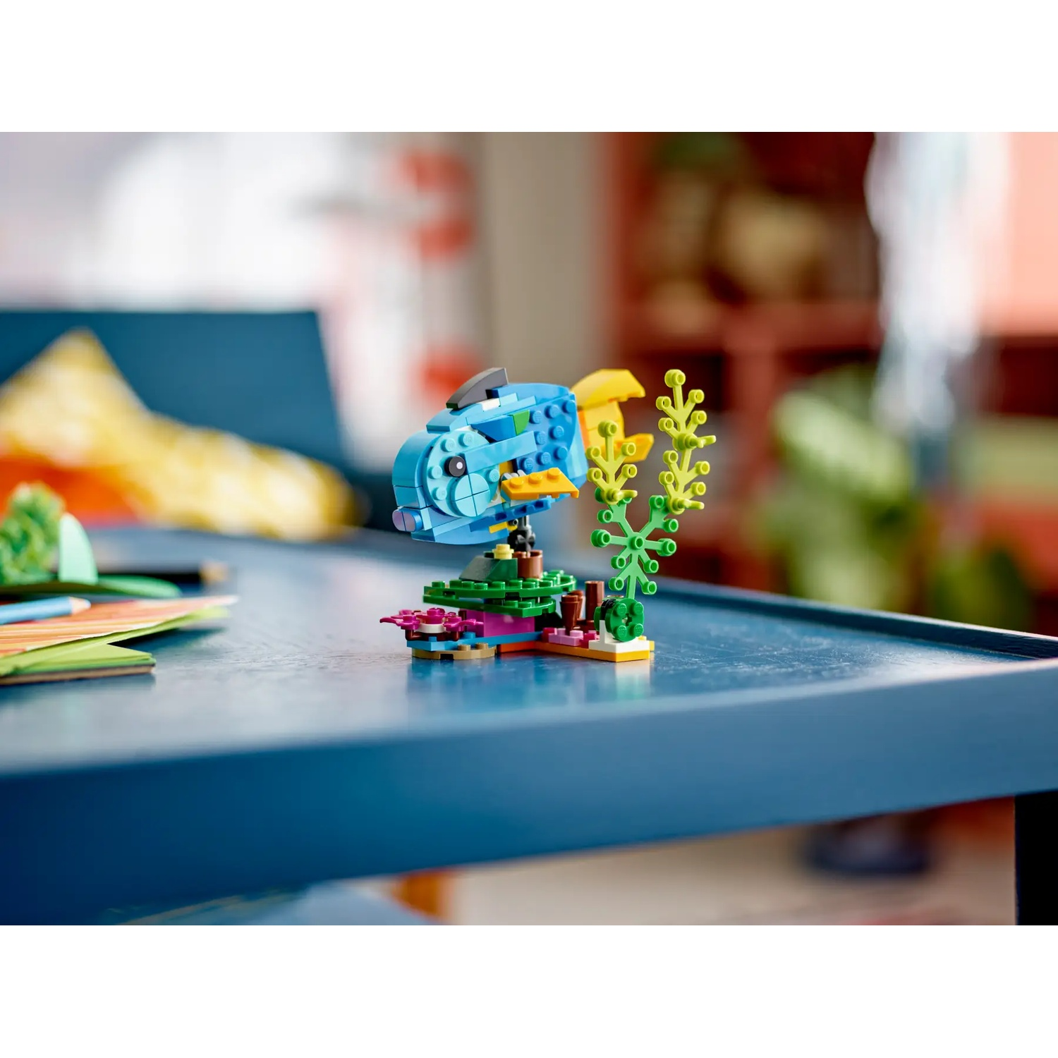 Een Lego-vis, blauw met gele vinnen, staat op een blauwe tafel. Groene plant en andere Lego-stukken zijn ook zichtbaar.