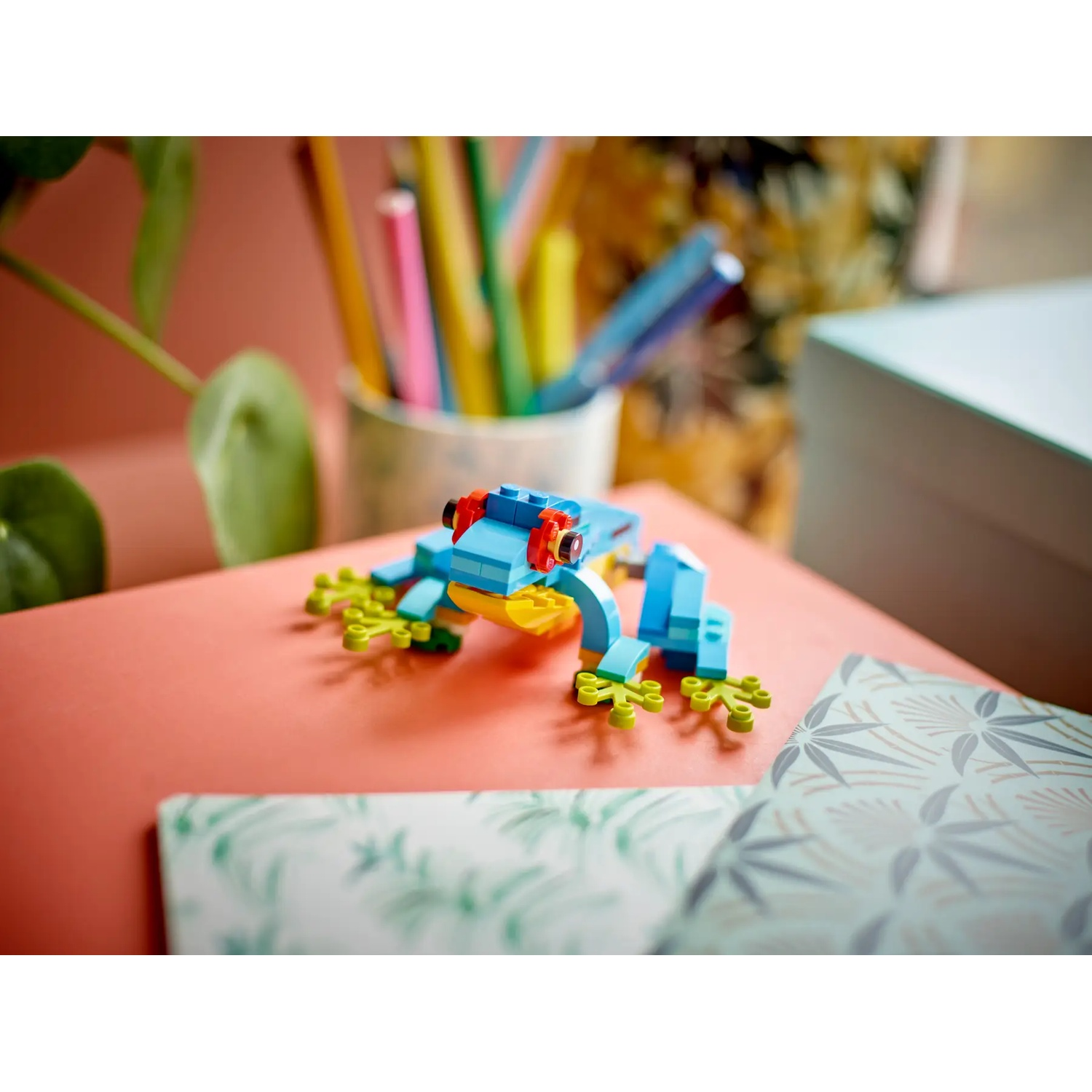 Een Lego-kikker gemaakt van blauwe, gele, rode en groene stenen. Hij zit op een roze oppervlak met briefpapier en een notitieblok met patroon op de achtergrond.