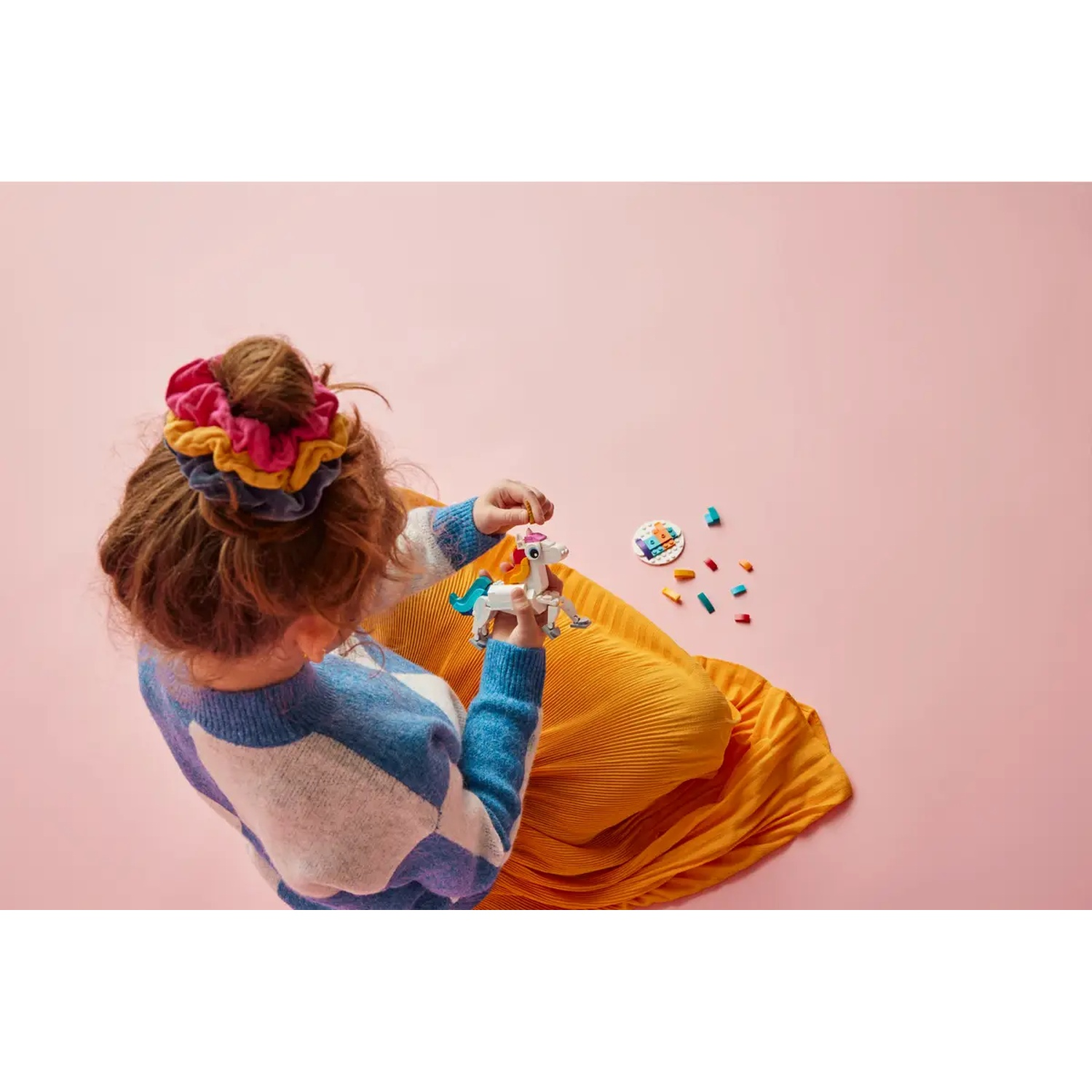 Une fille avec un chouchou tient une licorne jouet. Elle porte un pull bleu et une jupe jaune. Des blocs colorés et un fond rose.