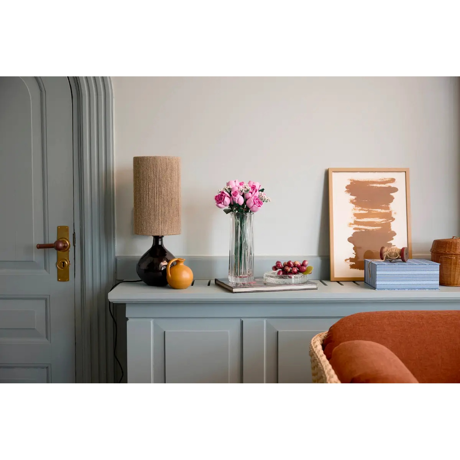 Een kamer met een grijze kast, een lamp, een vaas roze bloemen en een fotolijst. Een blauwe doos en een mand staan op de kast.