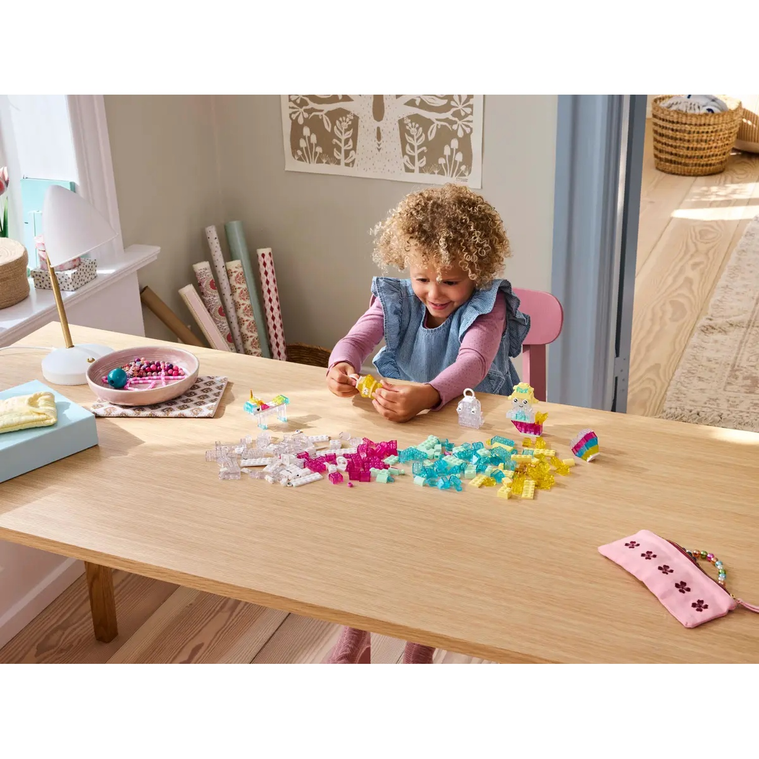 Une fille est assise à une table et joue avec des blocs colorés. Divers jouets et articles de bricolage sont sur la table. La pièce a une palette de couleurs neutres.