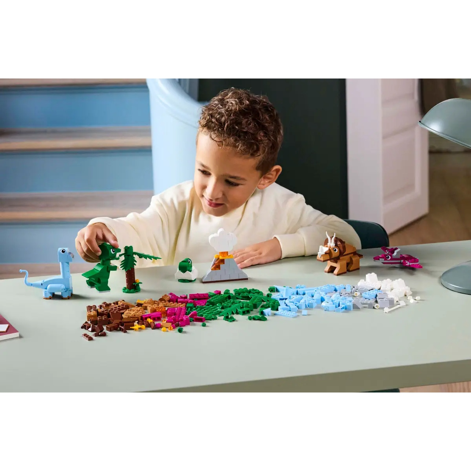 Un garçon joue avec des dinosaures Lego, un volcan et d'autres blocs sur une table. Différentes couleurs de blocs sont dispersées.